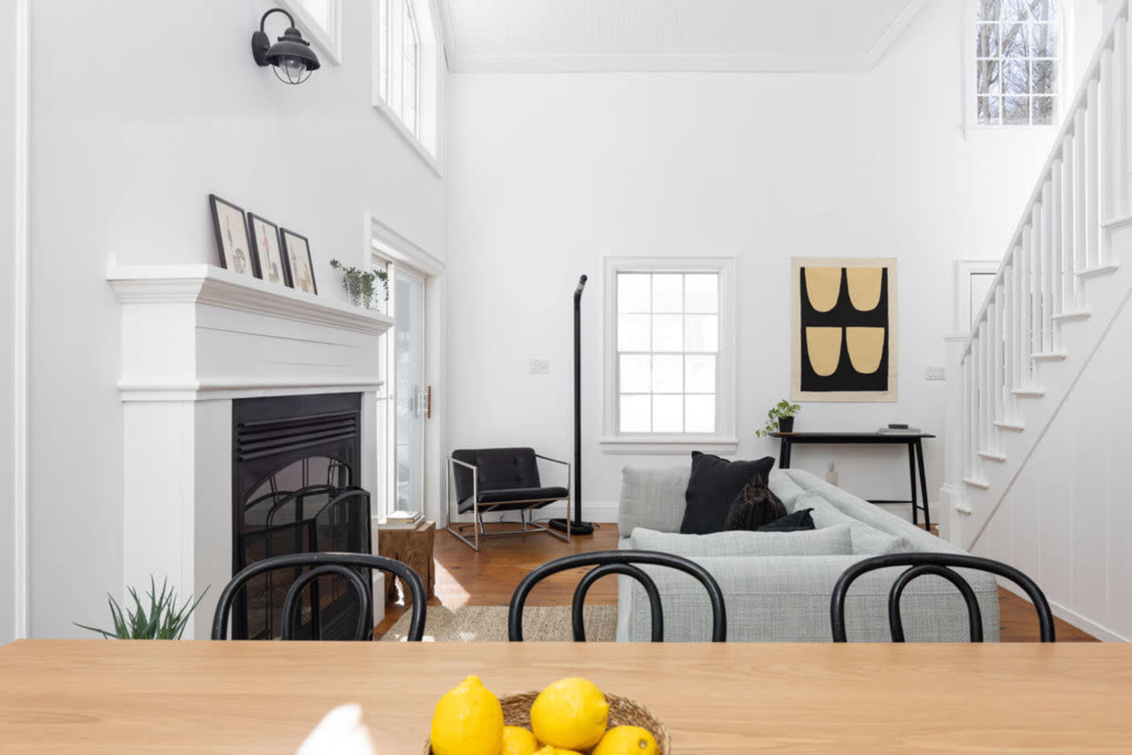 The image shows a bright, modern living room with a sofa, a fireplace, and large windows, alongside a dining area featuring a table with yellow lemons.
