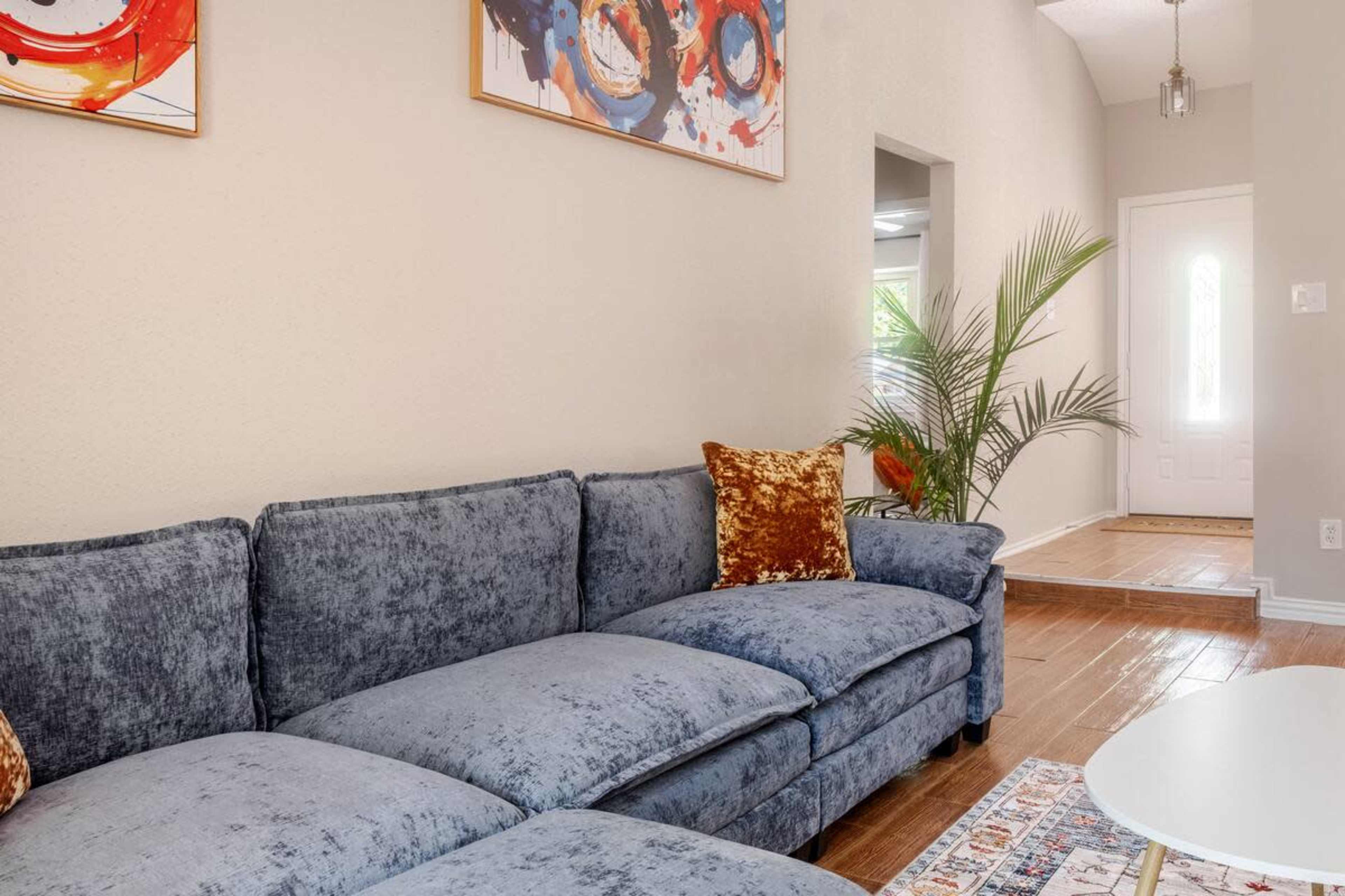 A blue sectional couch is positioned in a brightly lit living room with a decorative plant and a front door visible in the background.