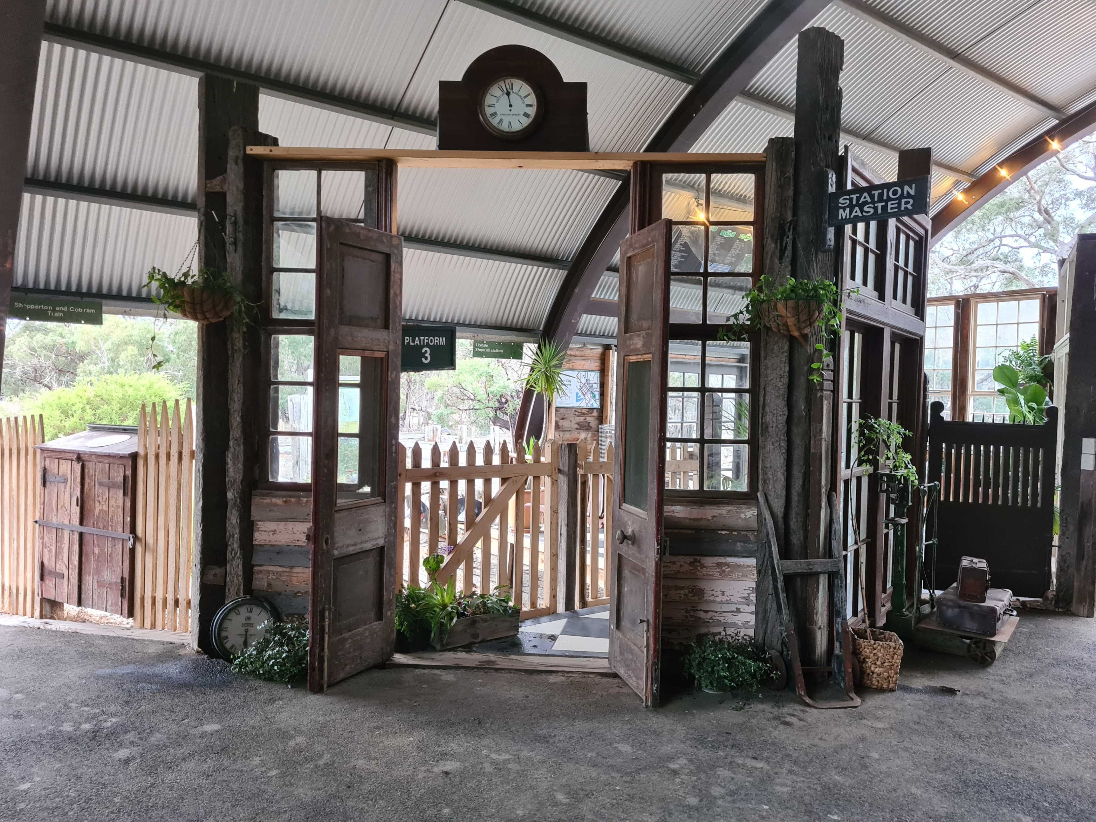 The image shows a rustic train station entrance with double doors, surrounded by wooden structures, a clock on the wall, and decorative plants.