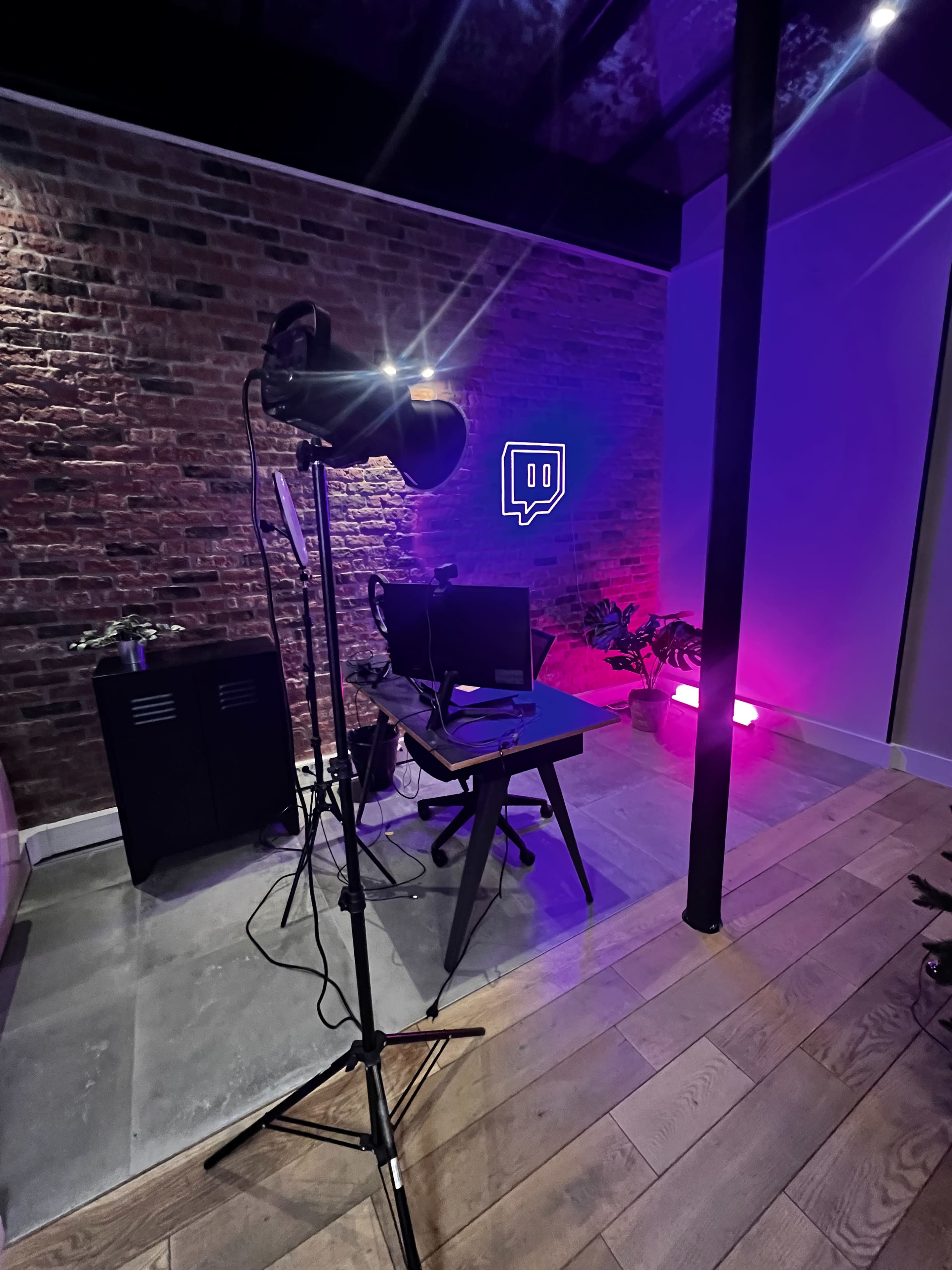 The image shows a modern workspace featuring a black desk with a computer setup, a lighting equipment stand, and a neon Twitch logo on a brick wall.