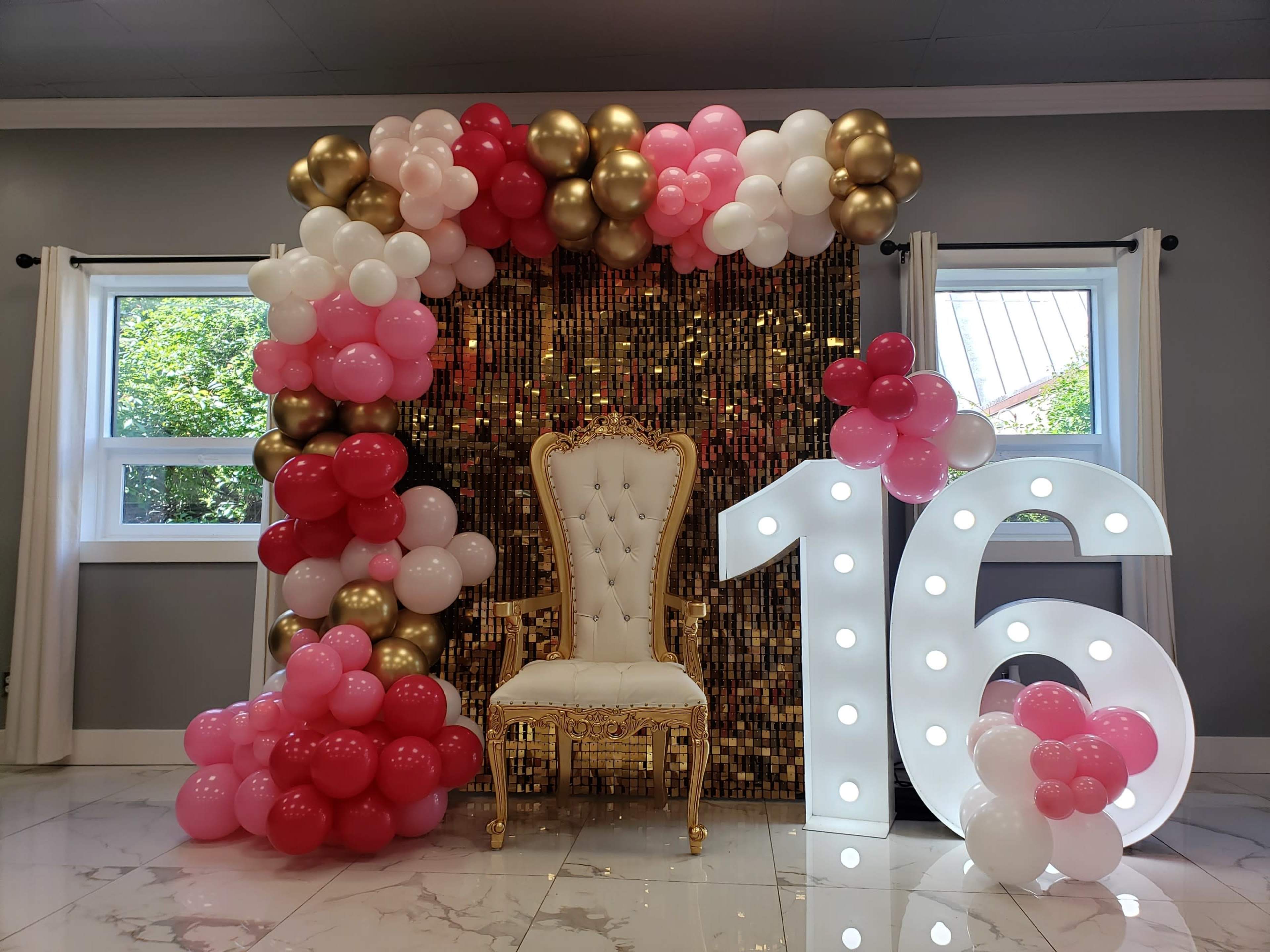 The image shows a decorative setup featuring a golden throne surrounded by a balloon arch in pink, white, and gold, with a large illuminated "16" nearby.