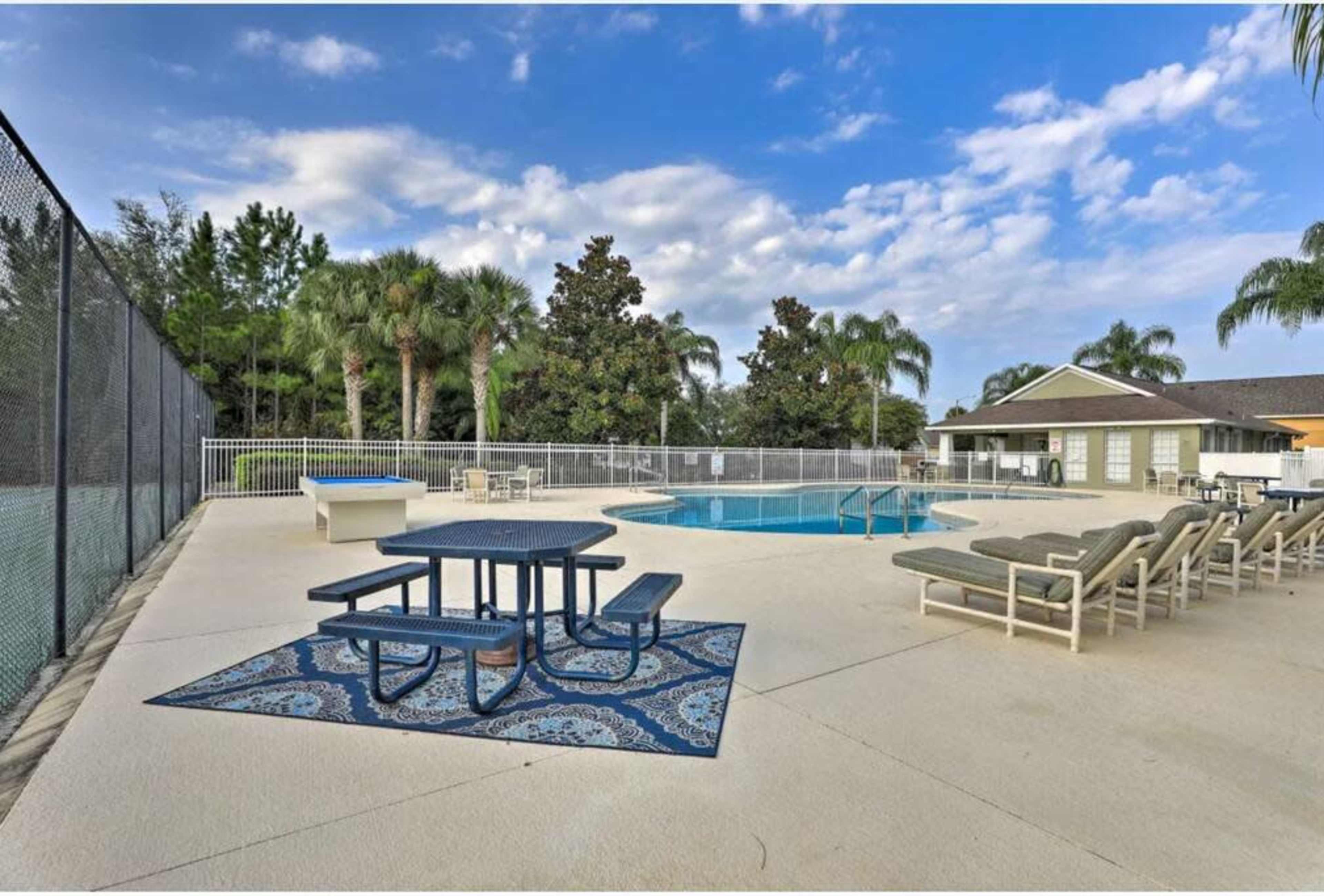 The image shows a poolside area with a picnic table and lounge chairs, surrounded by palm trees and a tennis court in the background.