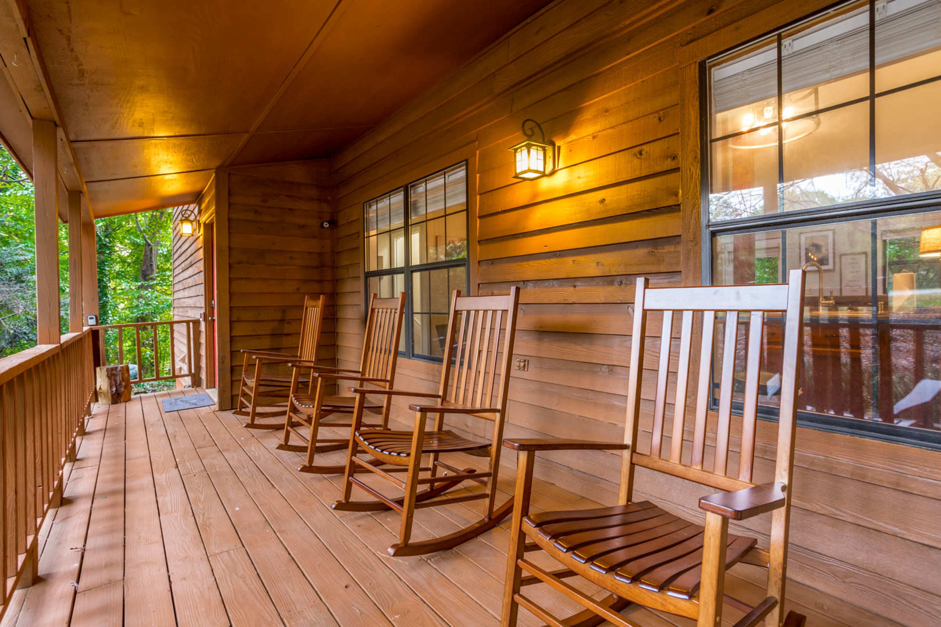 A wooden porch featuring four rocking chairs, with walls and beams made of brown-stained wood and lit by wall-mounted lamps.