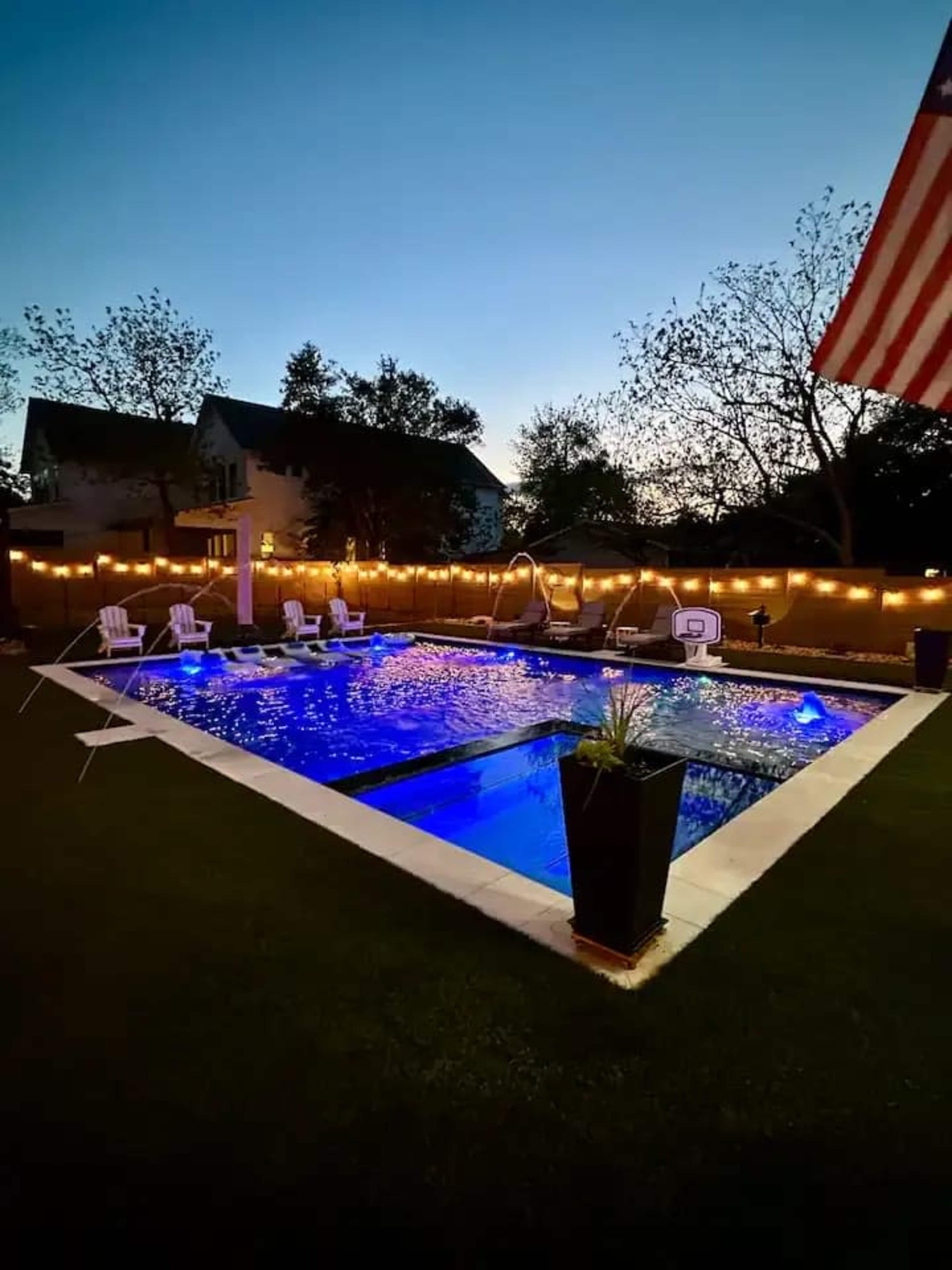The image shows a well-lit backyard pool area with a rectangular swimming pool surrounded by lounge chairs, illuminated by string lights and an American flag in the foreground.