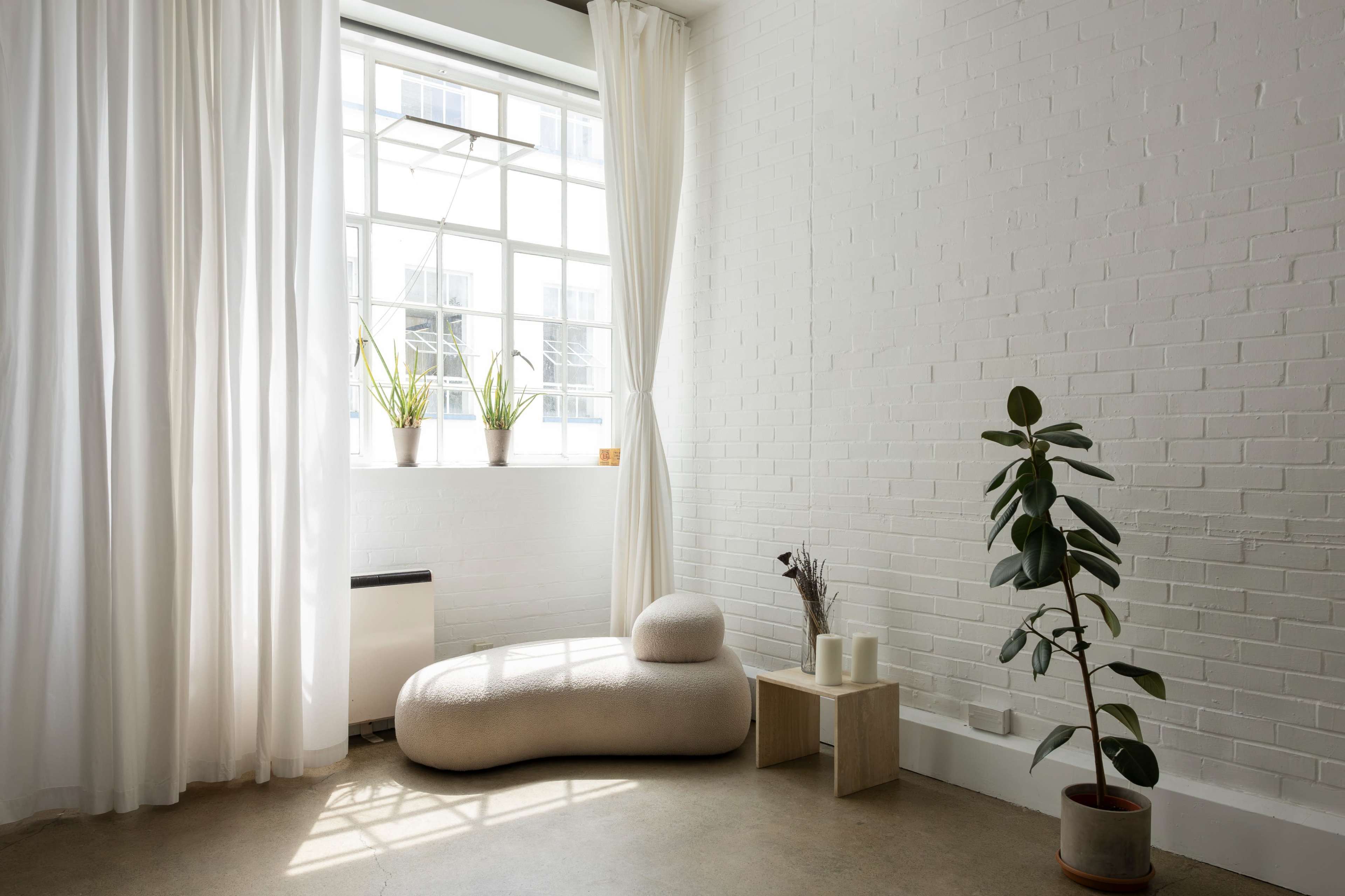 The image features a minimalist interior with a curved, beige seating area, white curtains, a large window, small potted plants, and a wooden side table.