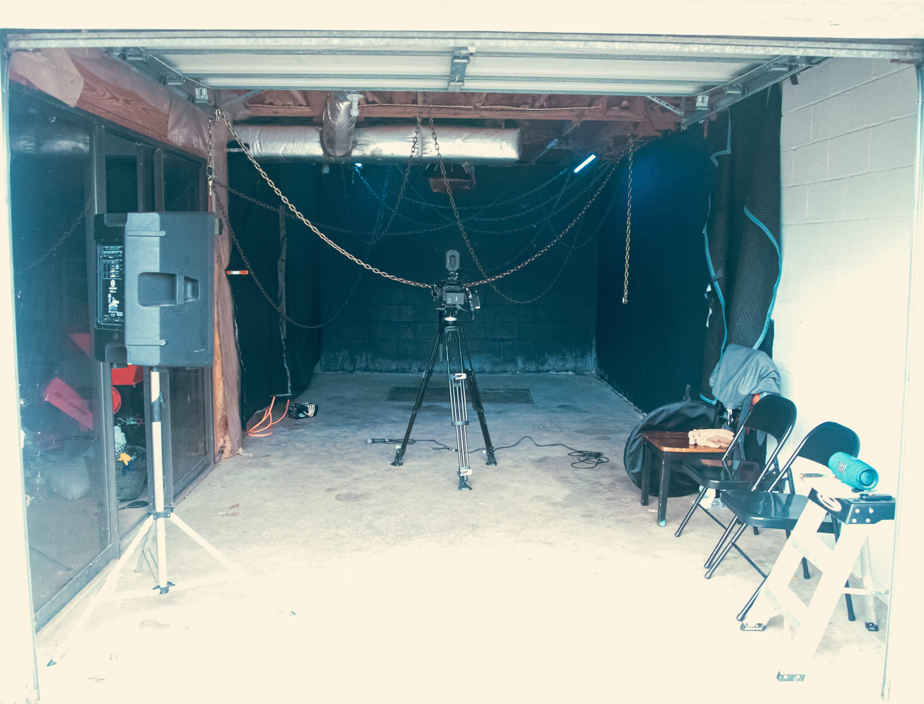 The image shows a vacant garage space with a camera setup on a tripod, a small table, and several chairs.