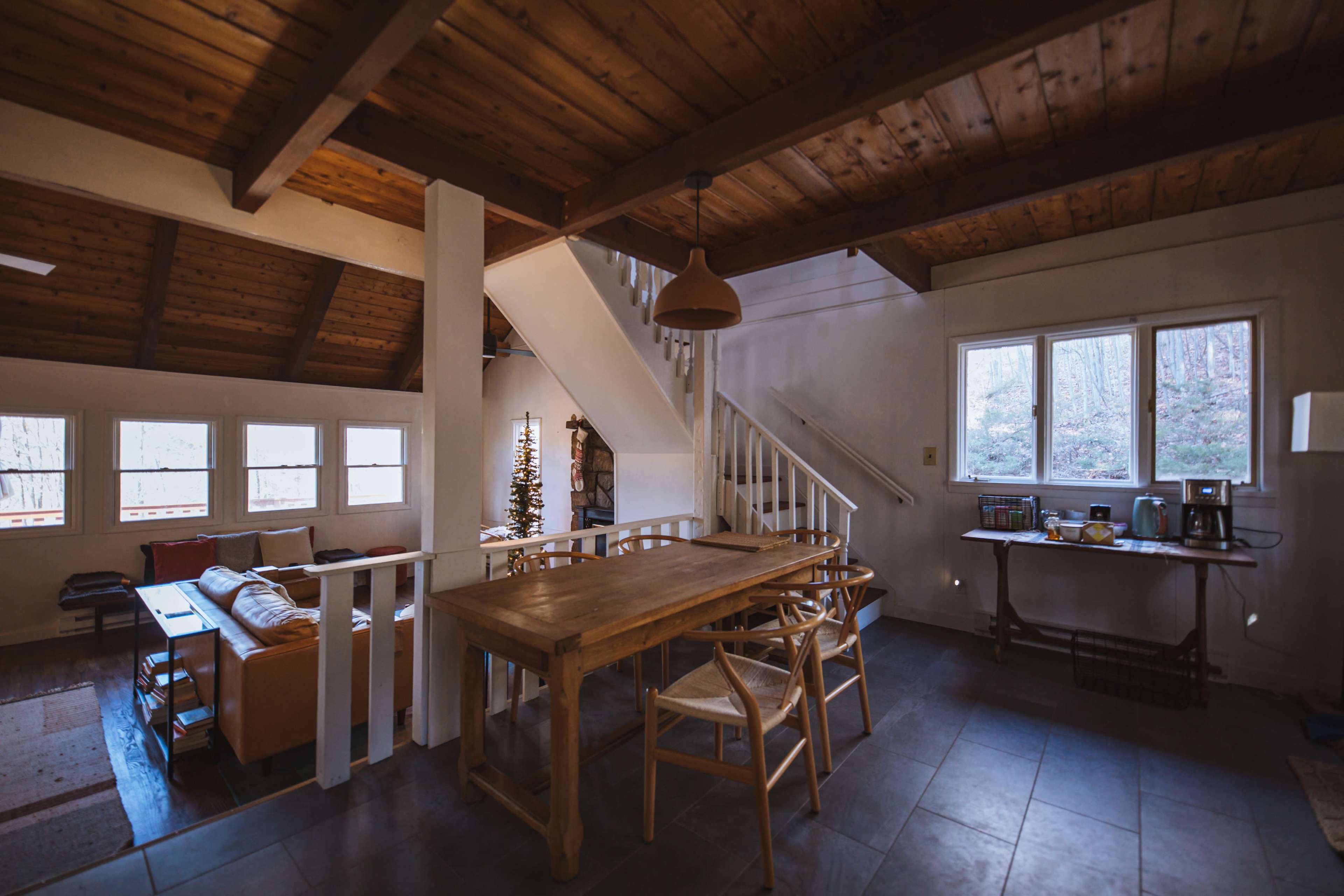 A cozy interior of a cabin featuring a wooden dining table, a staircase leading to an upper level, and a living area with a couch near large windows.