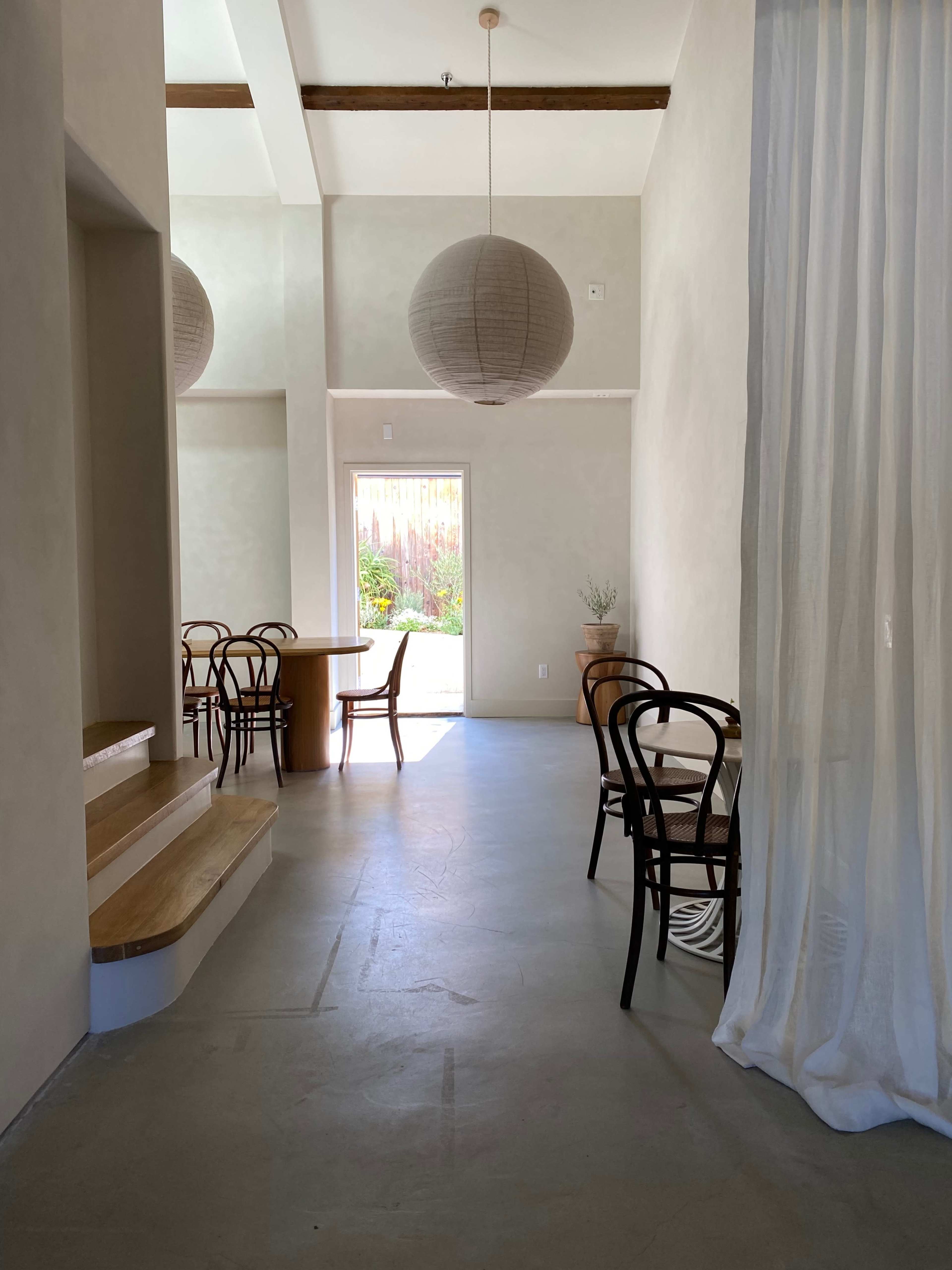 A minimalist interior with a wooden dining table, bentwood chairs, a doorway leading to a garden, and hanging paper lanterns.