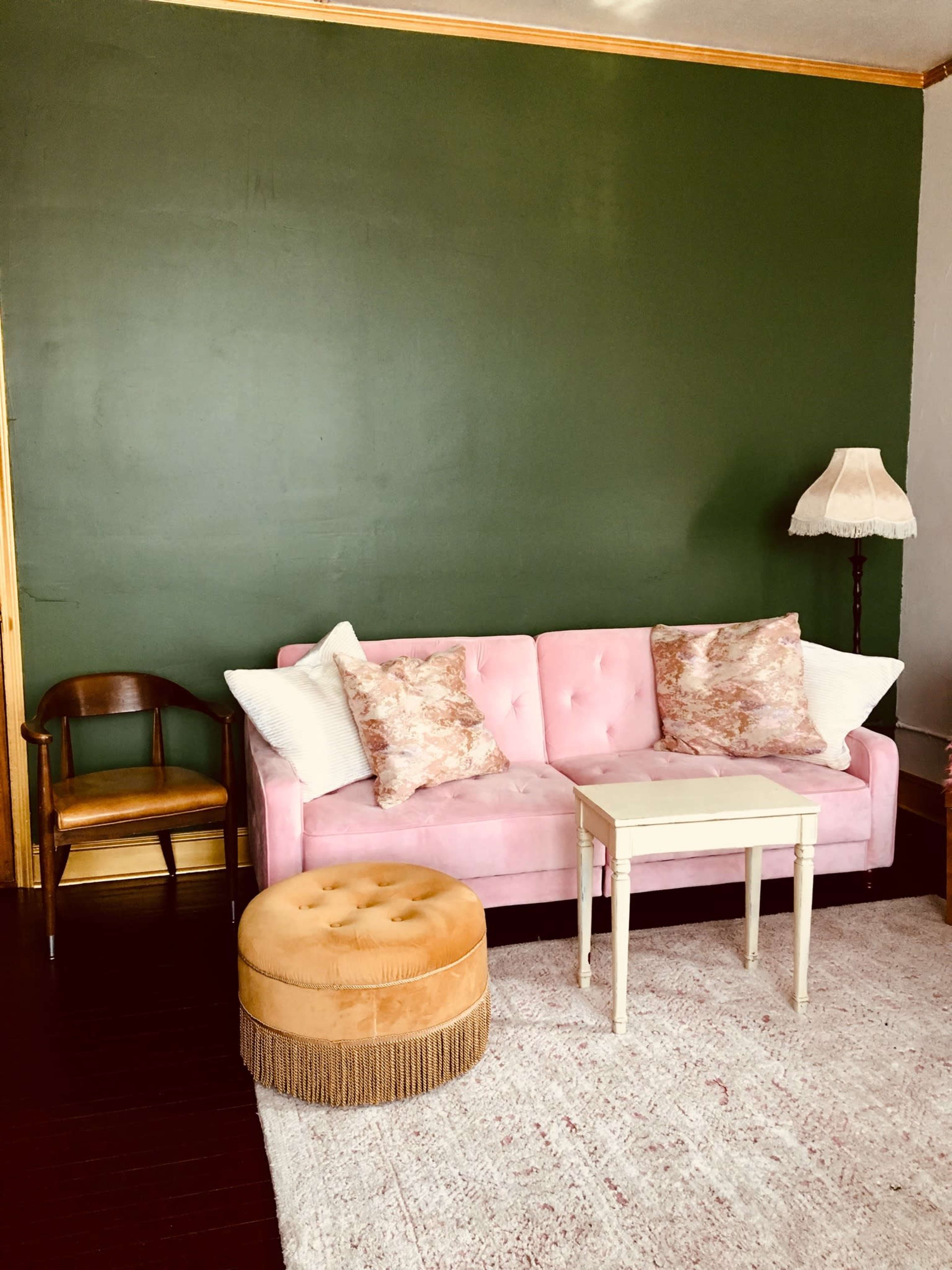 A pink velvet sofa with patterned pillows is positioned in front of a dark green wall, accompanied by a small white side table, a round ottoman, and a wooden chair.