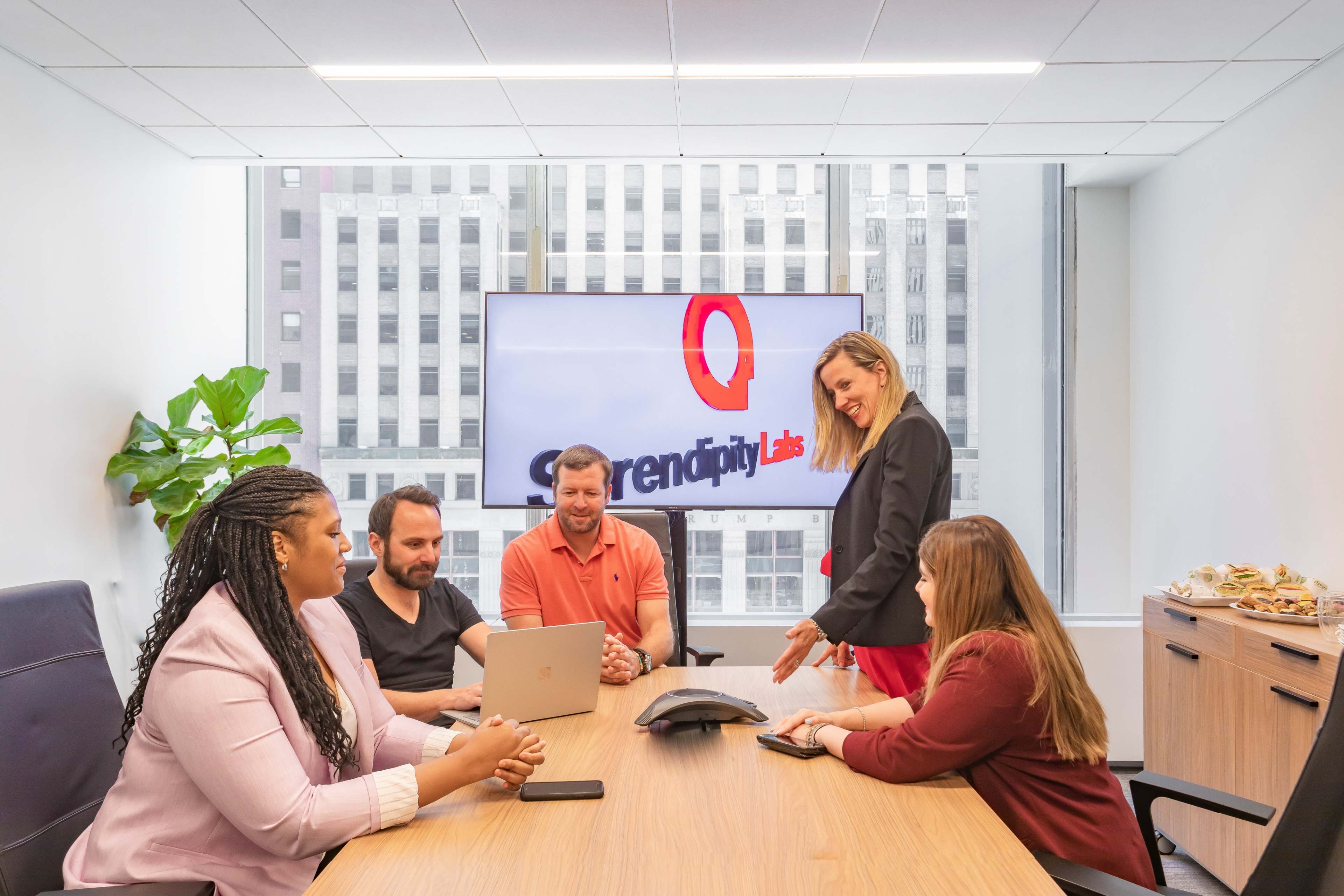 A group of five professionals is seated around a conference table in a modern office, engaged in a meeting with a large screen displaying the Serendipity Labs logo in the background.