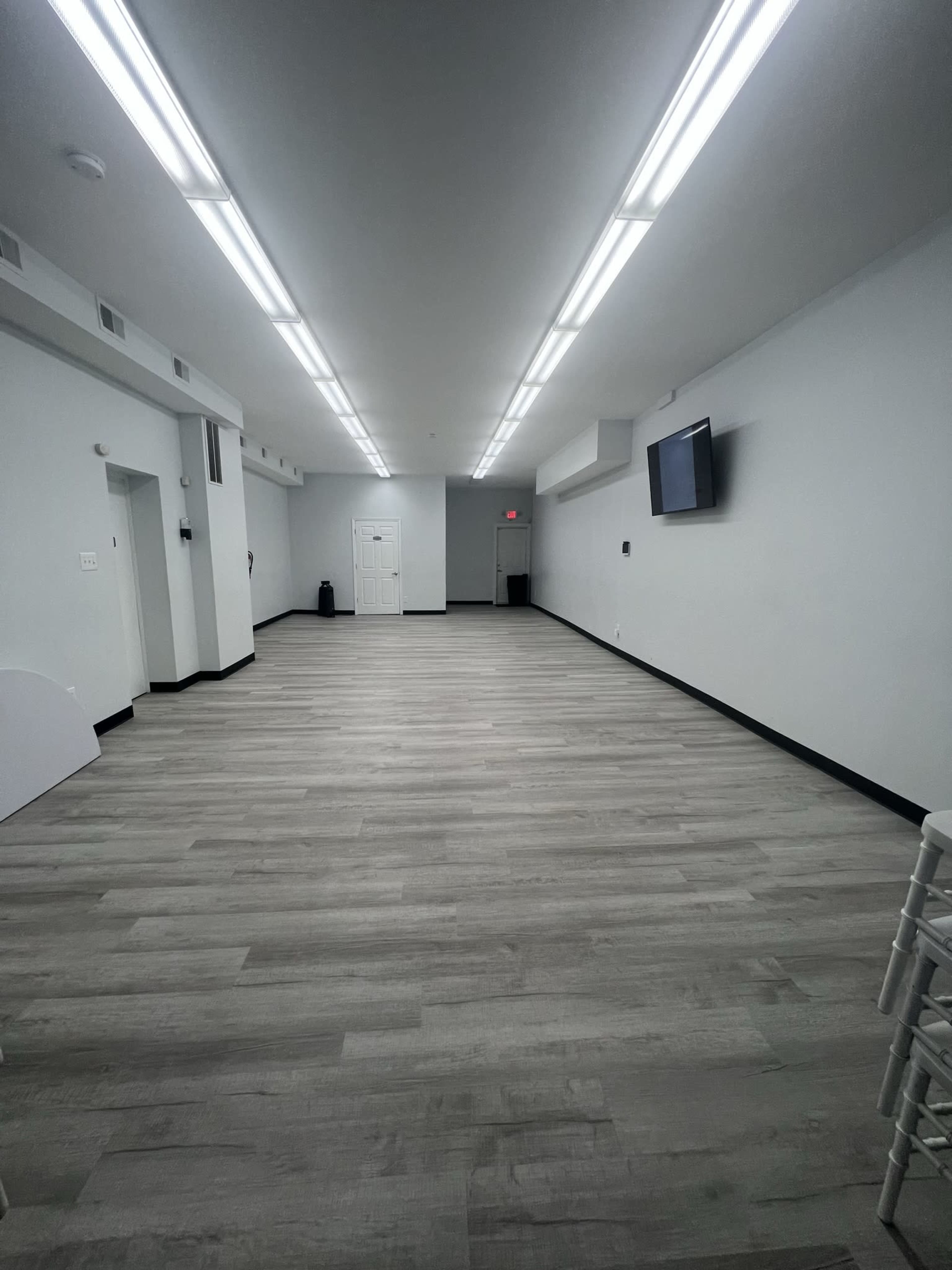 The image shows a spacious, empty room with gray wooden flooring, white walls, and bright overhead lights.