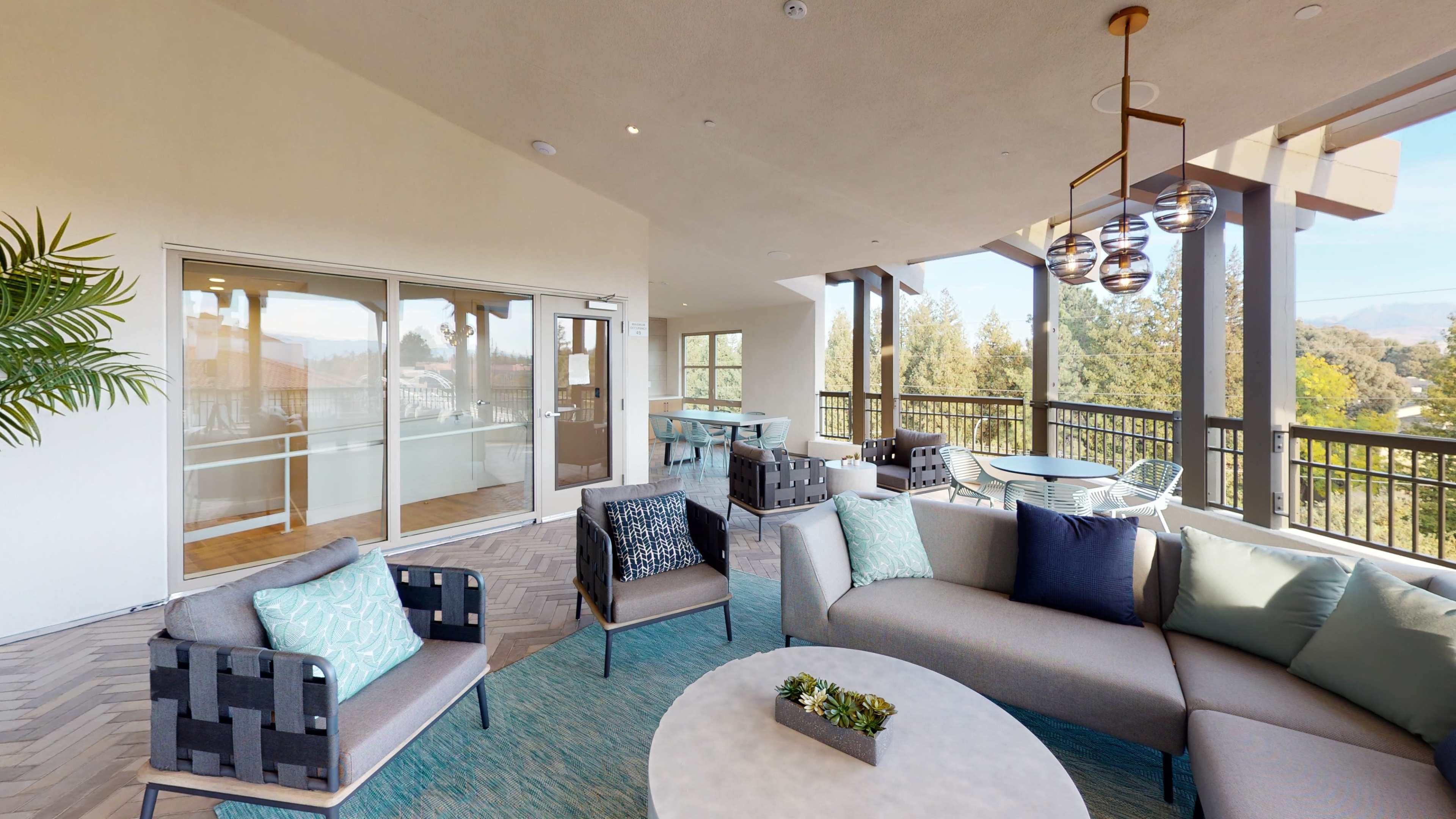 The image shows a modern patio space featuring seating arrangements, a circular table, and large glass doors leading to an outdoor area.