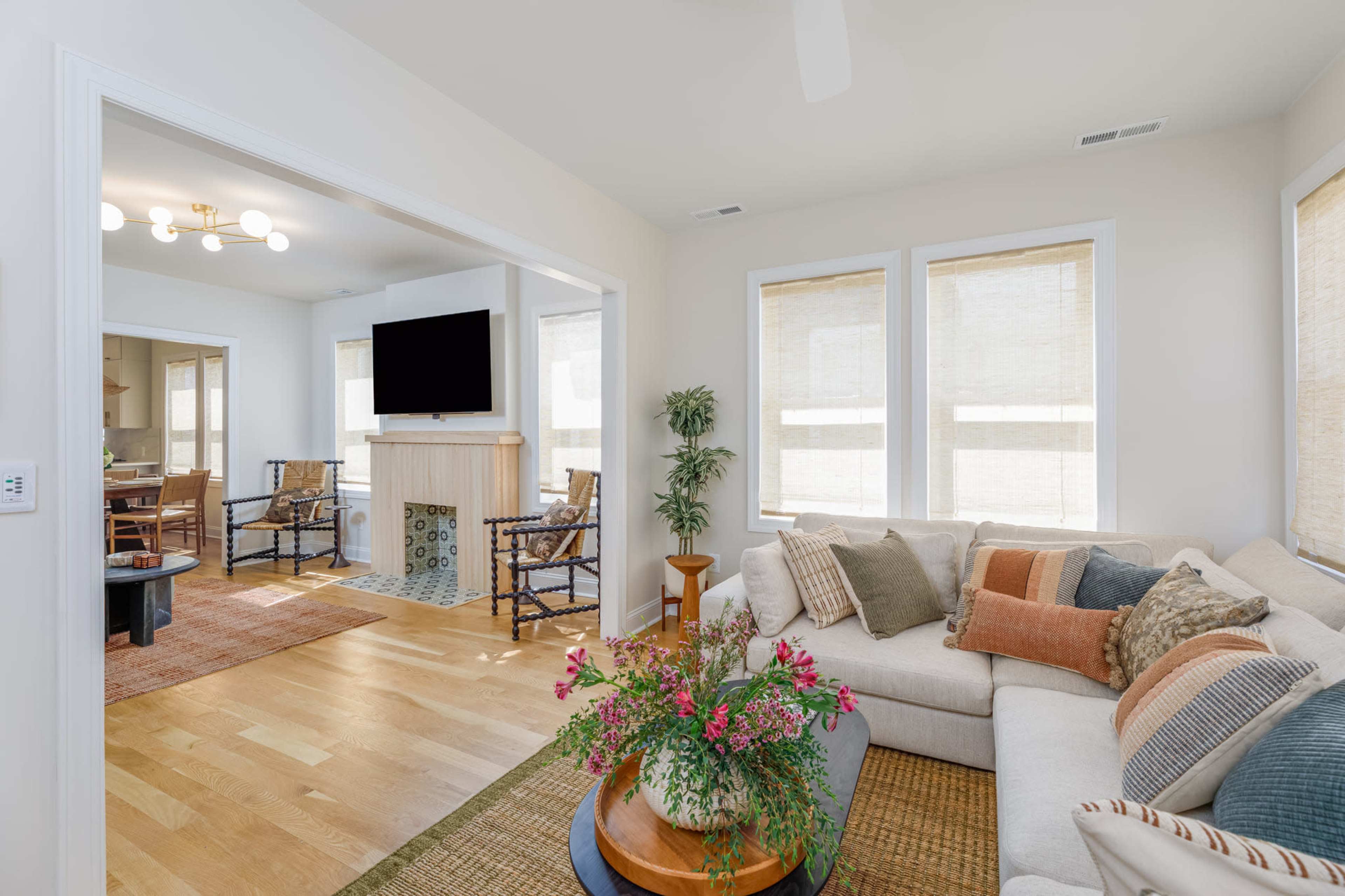A bright living room features a large sectional sofa, a coffee table with flowers, and windows adorned with shades, leading into a dining area with additional seating.