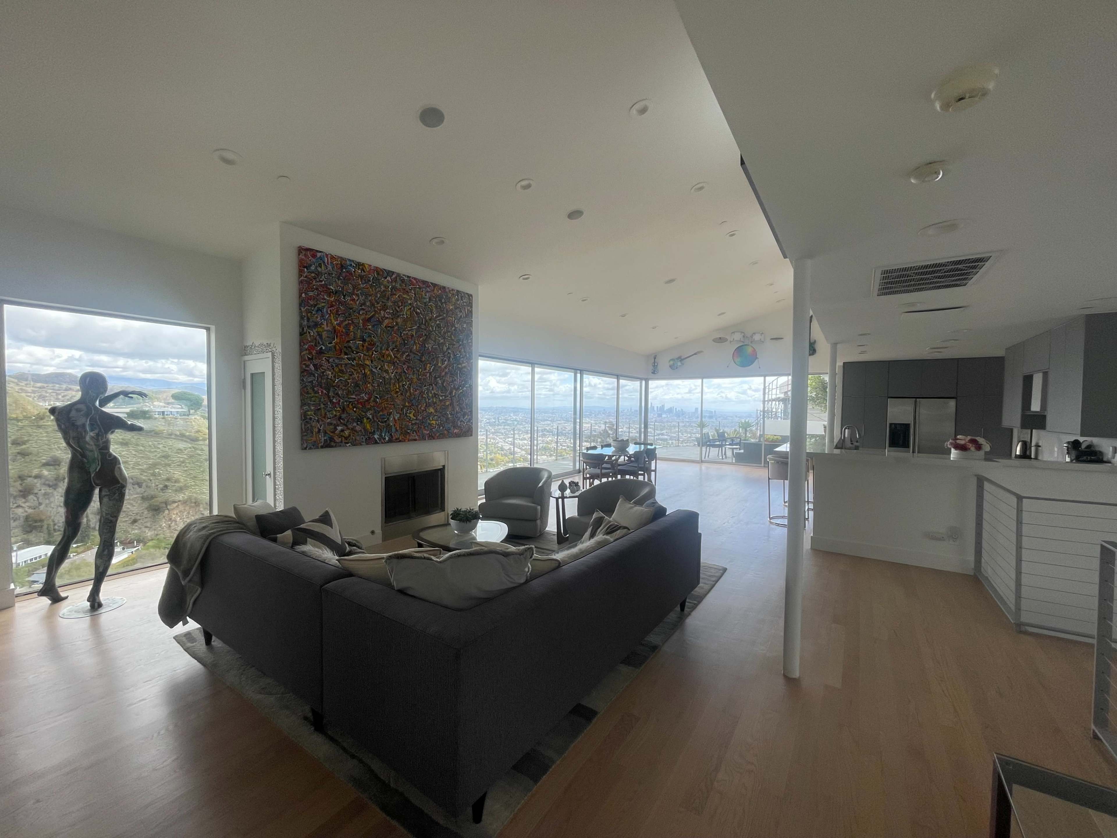 The image shows a modern, open-plan living space featuring a gray sofa, a large piece of abstract art on the wall, and expansive windows offering a view of the surrounding landscape.