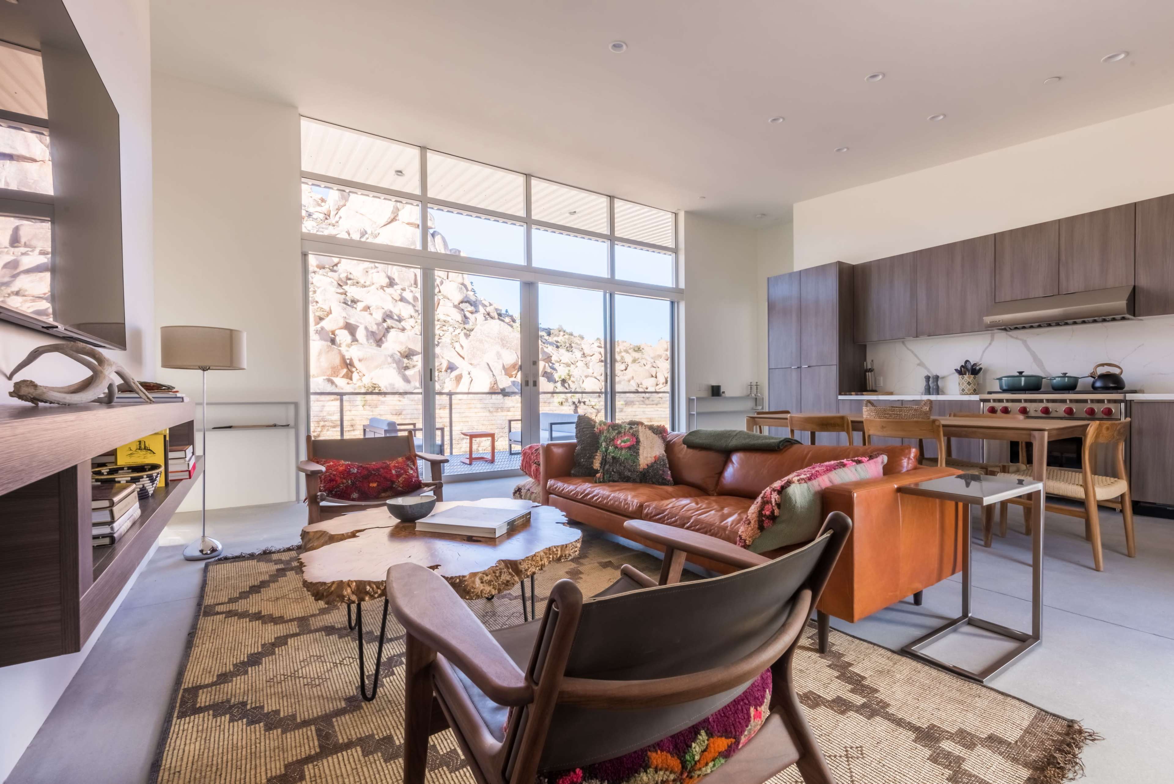 The image shows a modern living space with large windows overlooking rocky landscape, featuring a leather sofa, a wooden coffee table, and a kitchen area with sleek cabinetry.