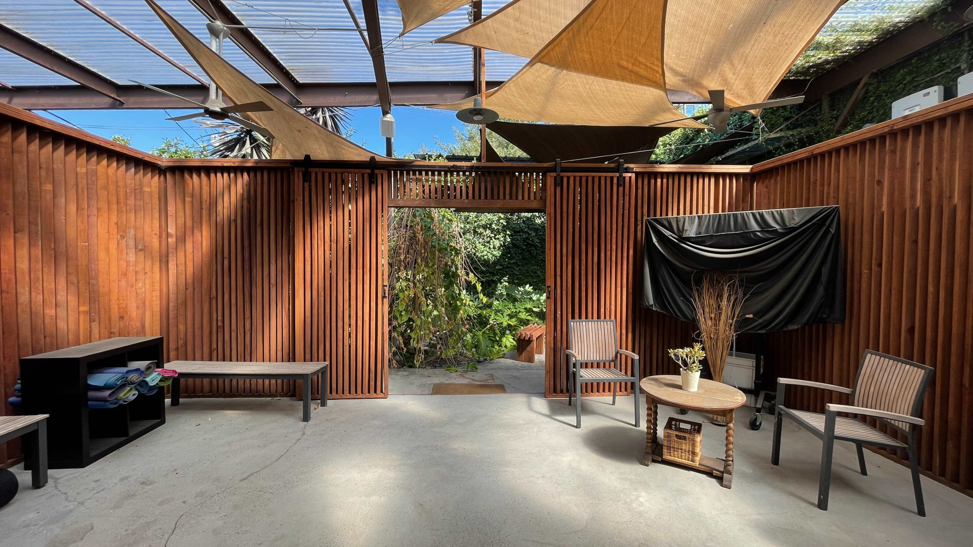 The image shows a wooden-walled outdoor space with a shaded area, featuring benches, chairs, and a small table, surrounded by lush greenery.