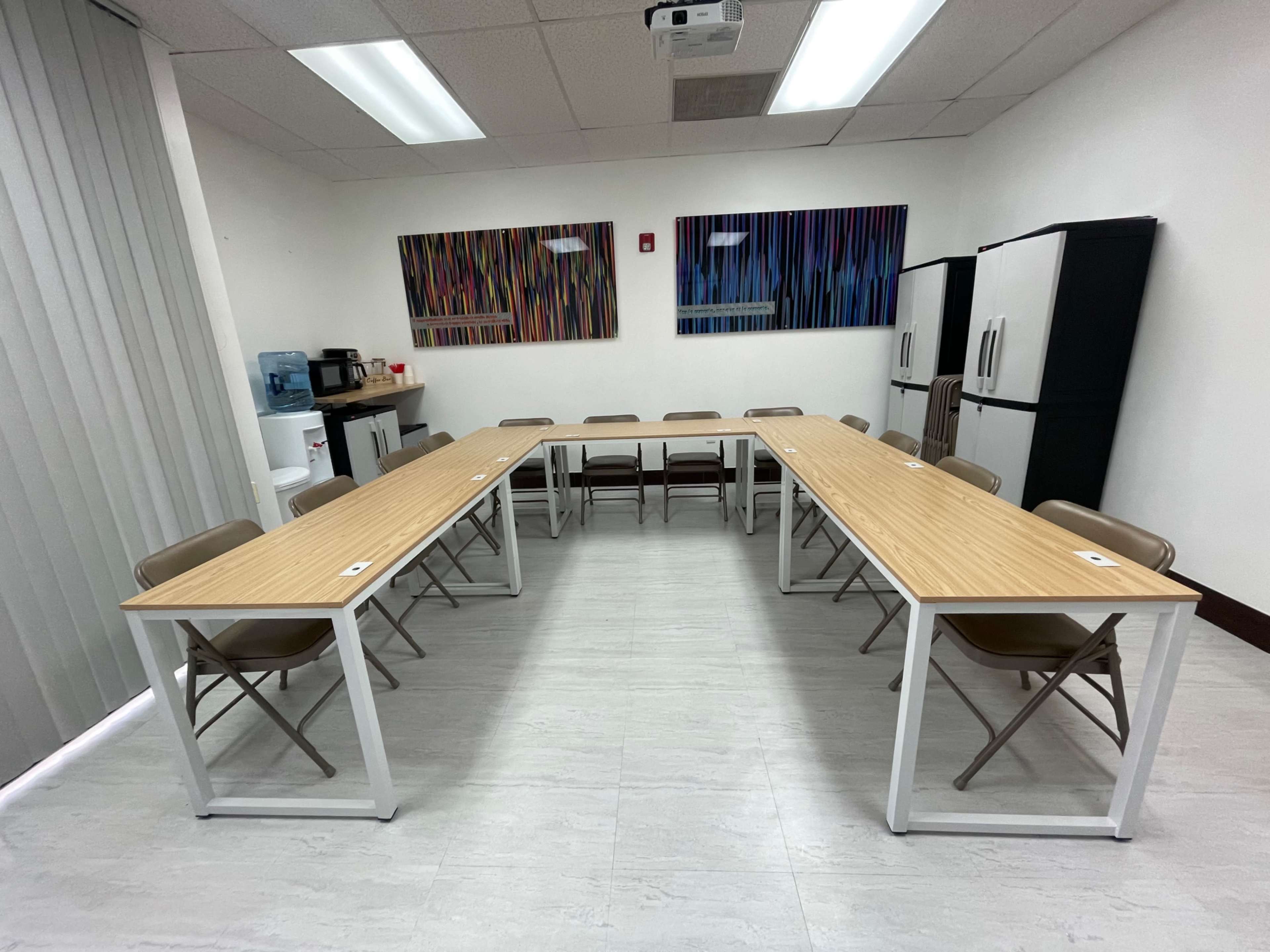 A meeting room is setup with two long tables arranged in a U-shape, surrounded by folding chairs, with a kitchen area visible in the background.