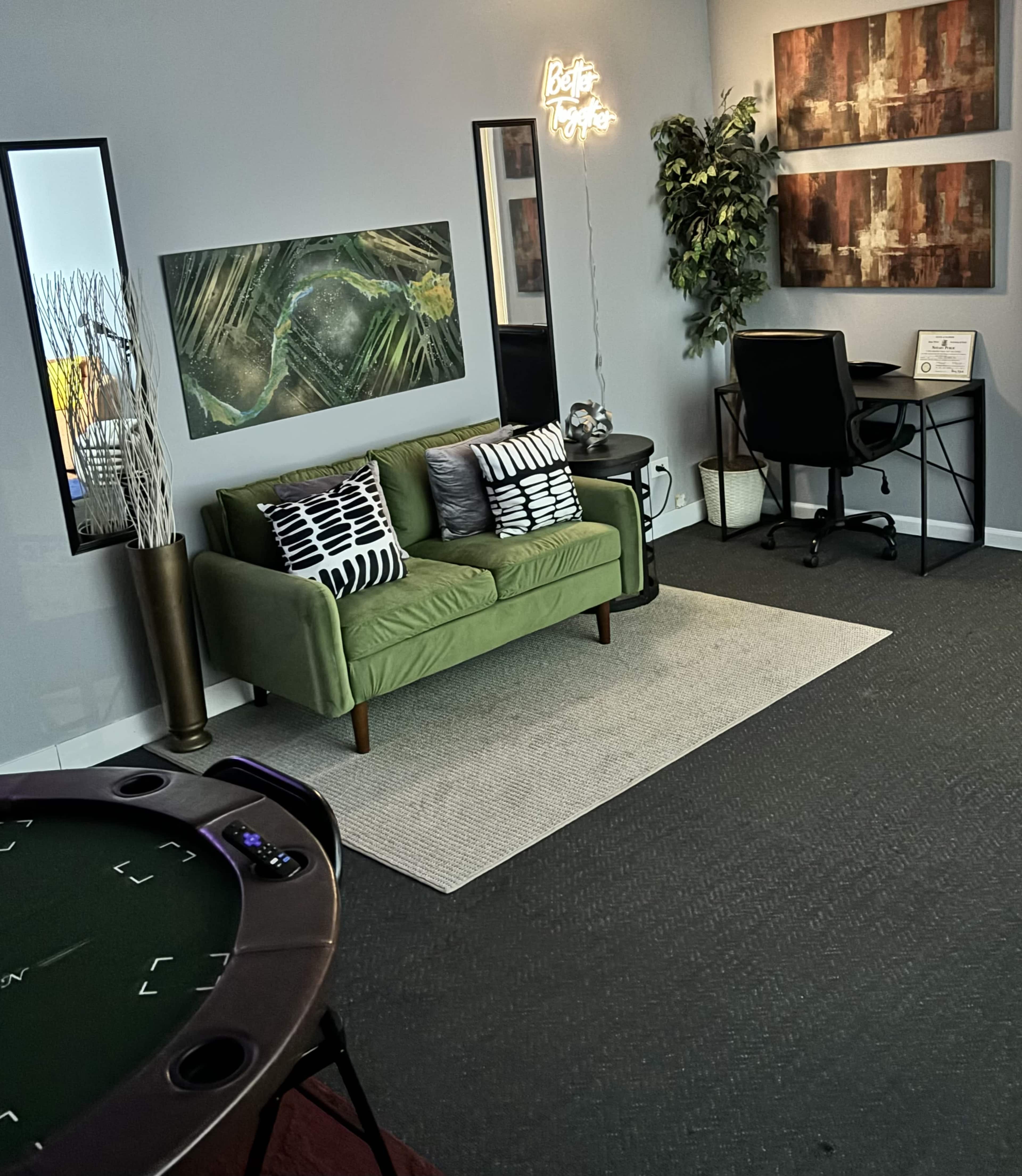 A modern office space featuring a green sofa, a desk with a chair, wall art, and a poker table in the foreground.