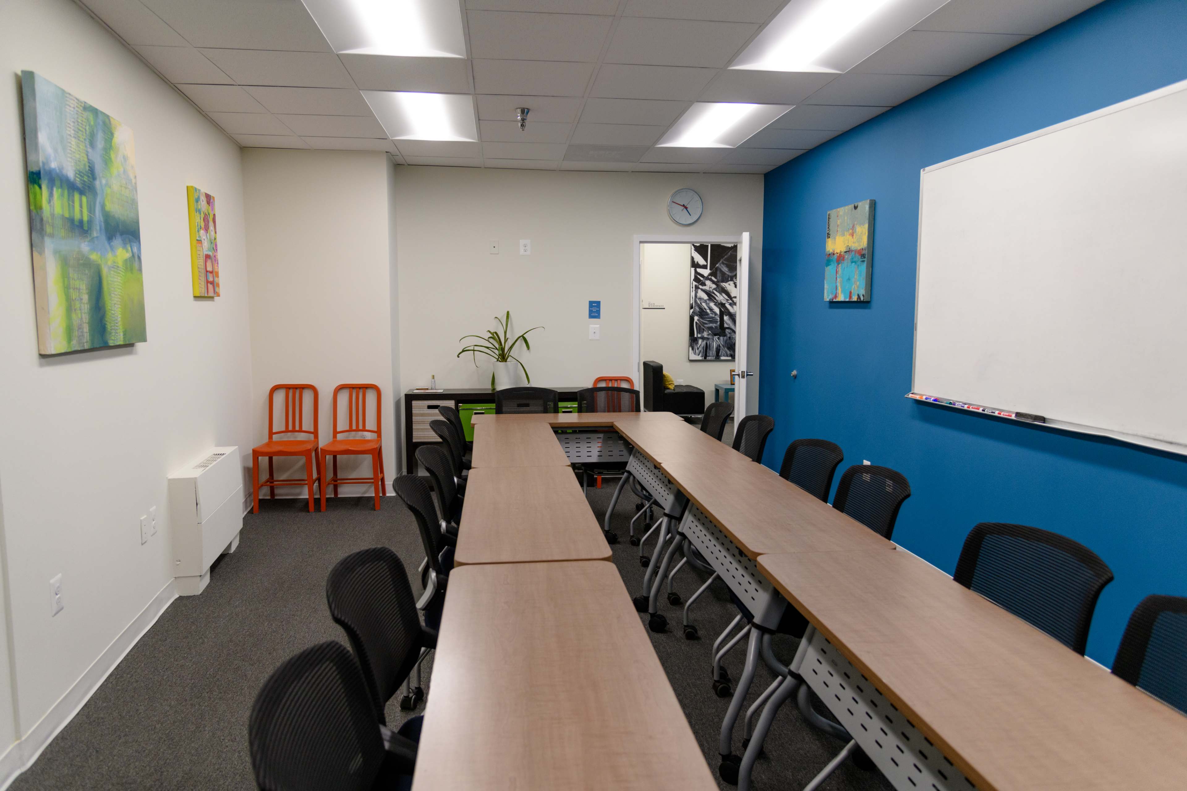 A conference room features a long table surrounded by black chairs, with colorful artwork on the walls and a whiteboard at one end.