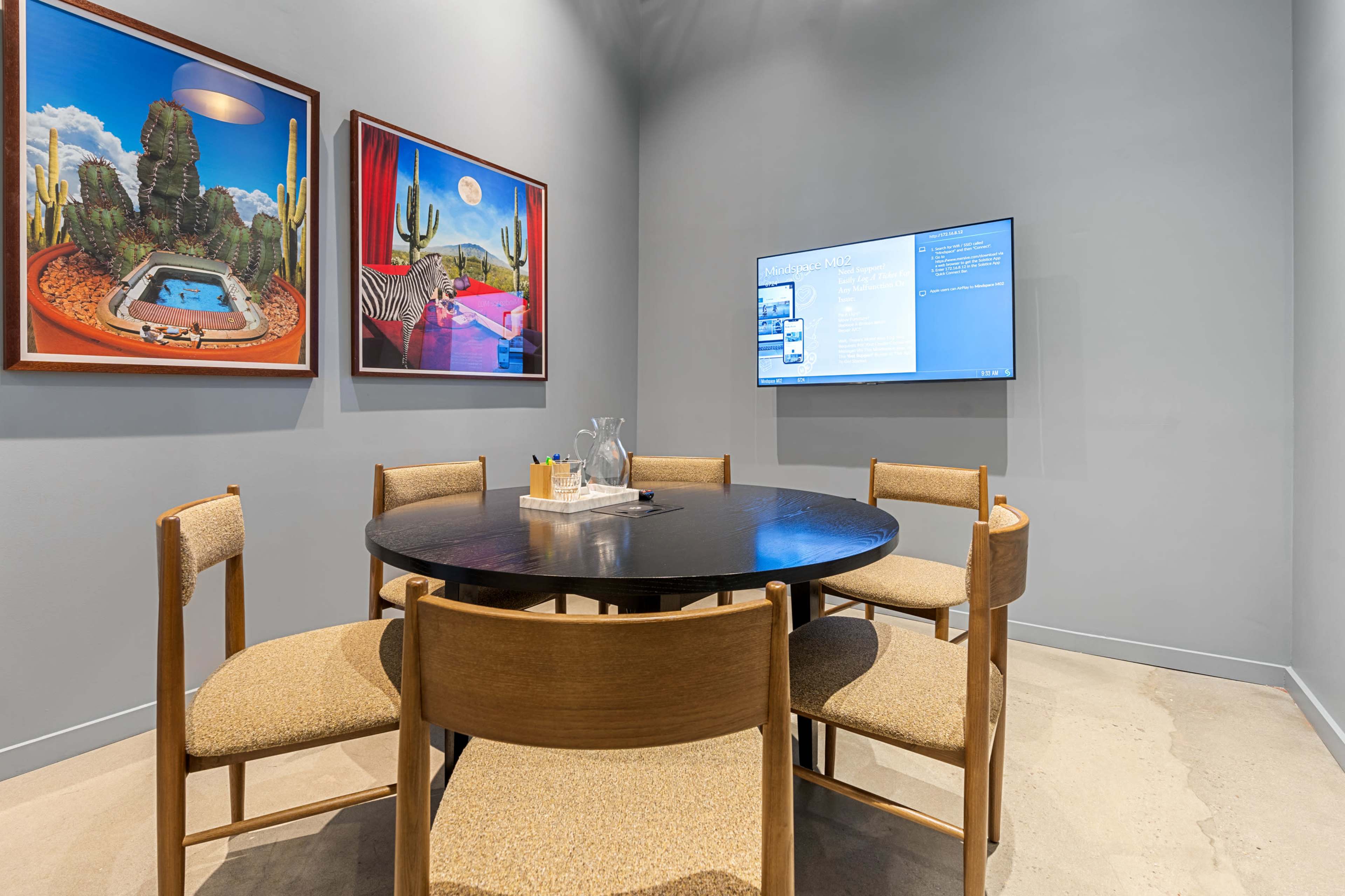 A small meeting room features a round black table surrounded by four wooden chairs, with two framed artworks on the wall and a television displaying information in the background.