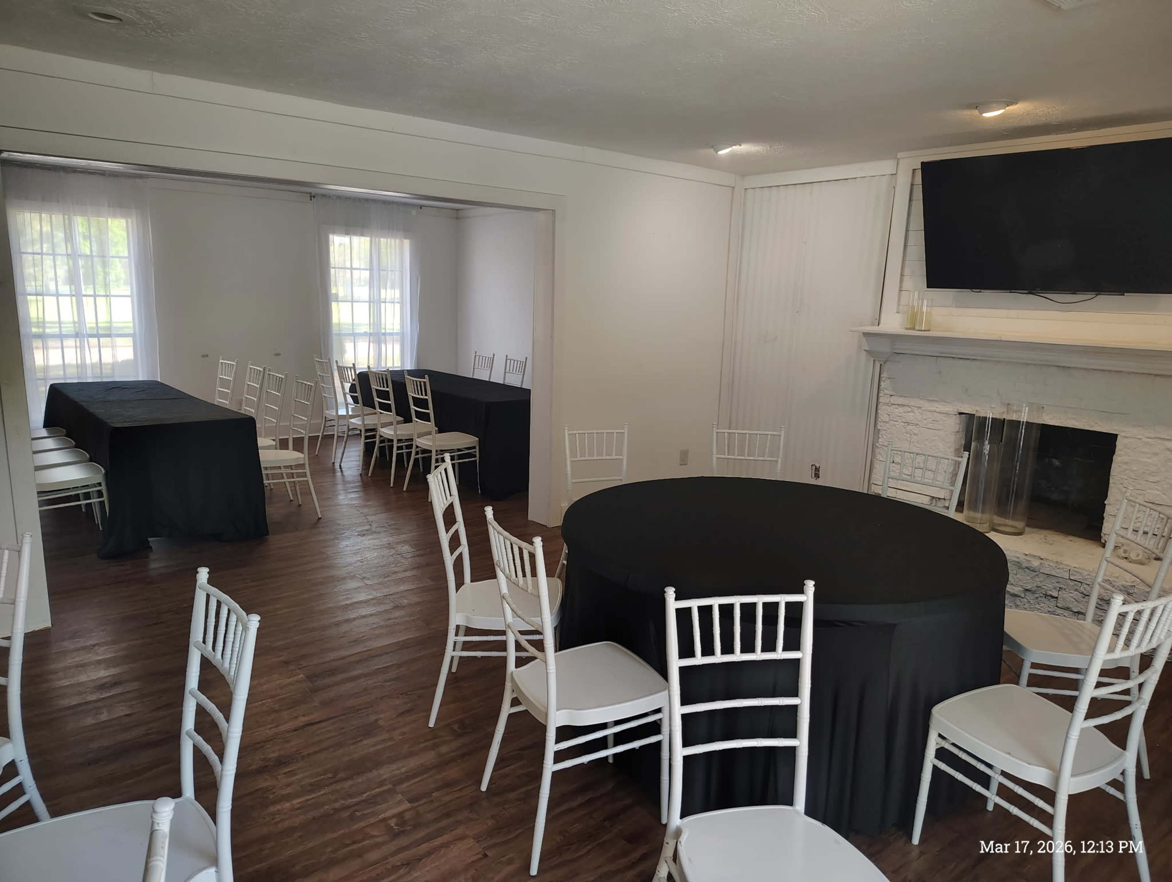 The image shows a spacious indoor room with two seating arrangements: a round table covered in a black tablecloth and several rectangular tables, all surrounded by white chairs.