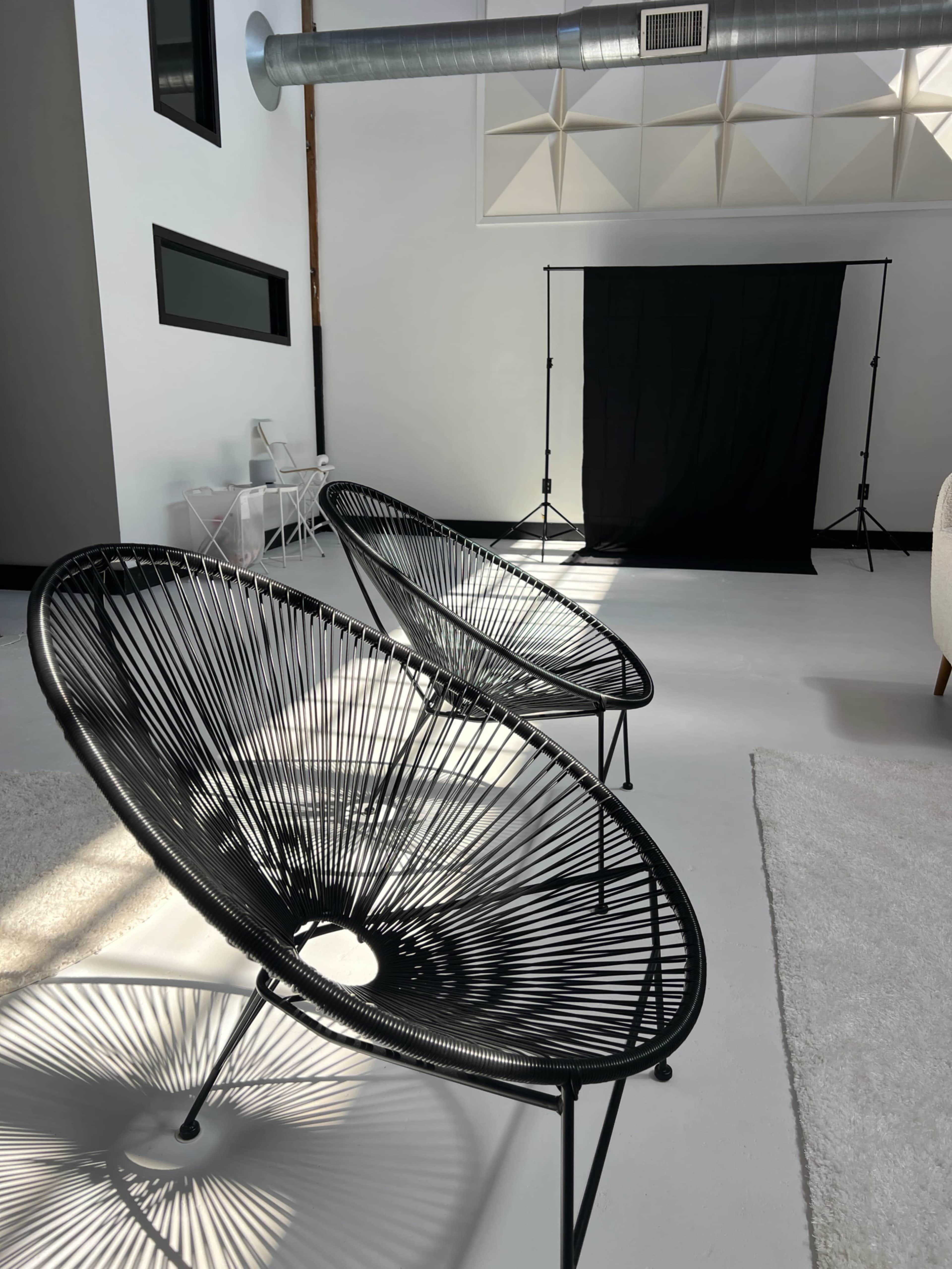 Two black wire chairs are positioned in a well-lit room with a white floor and a black backdrop set up for photography.
