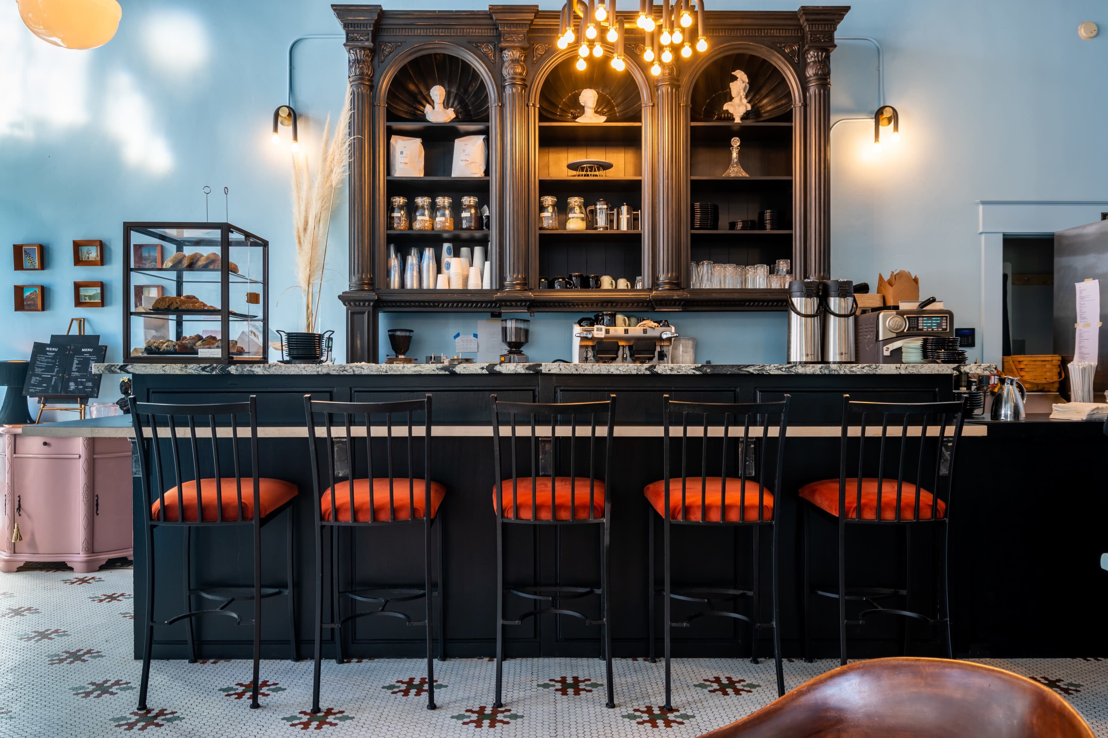 A stylish café bar features dark wood cabinetry, a marble countertop, and orange-cushioned stools lined up in front of it.