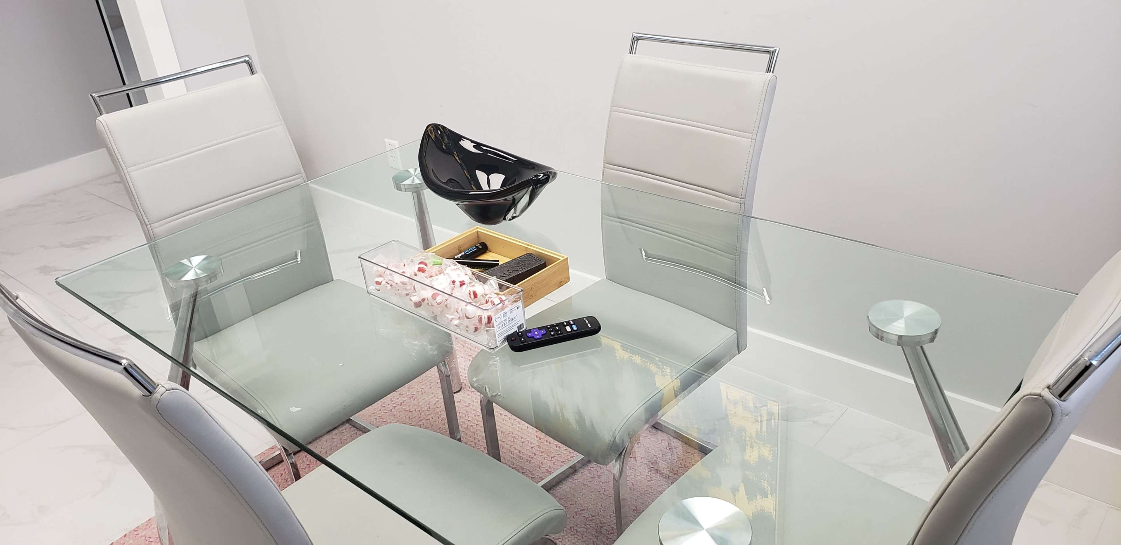 A glass dining table with four grey chairs and decorative items on top is positioned in a well-lit room.
