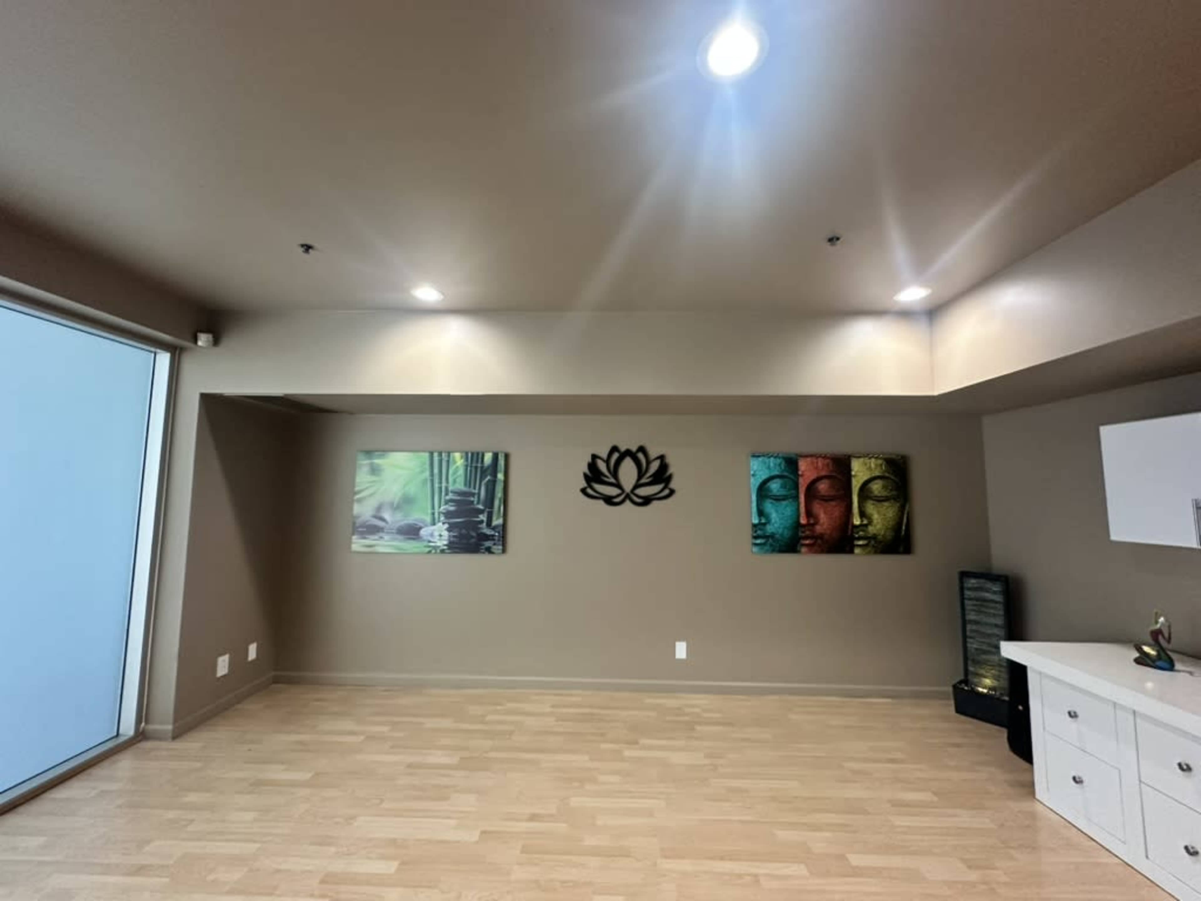 The image shows a spacious, well-lit room with wooden flooring, featuring two framed artworks of faces and a lotus design on the wall, along with a small water feature in the corner.