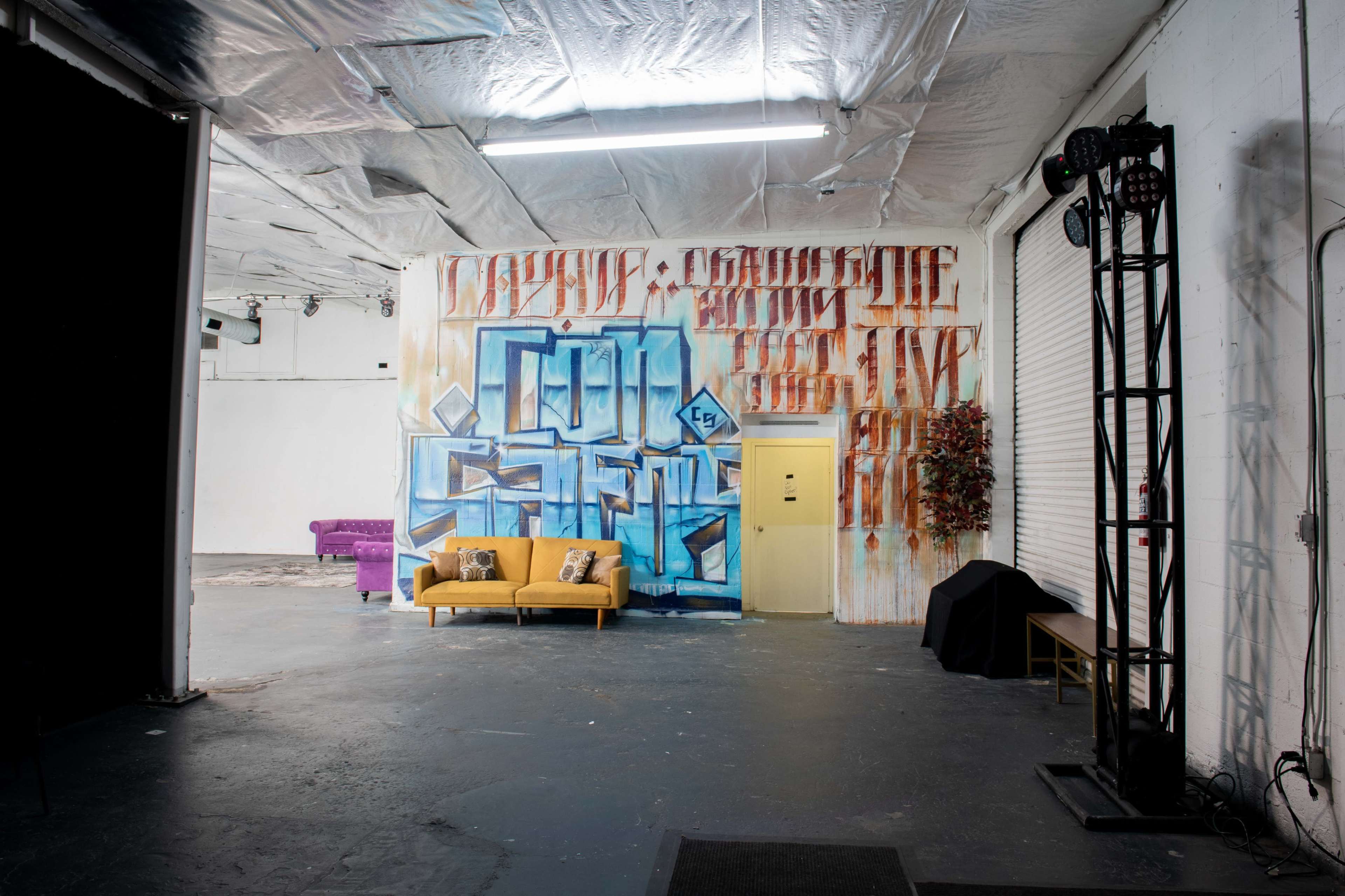 A spacious room with a yellow couch in front of a colorful graffiti wall, accompanied by a closed door and some stage lighting equipment.