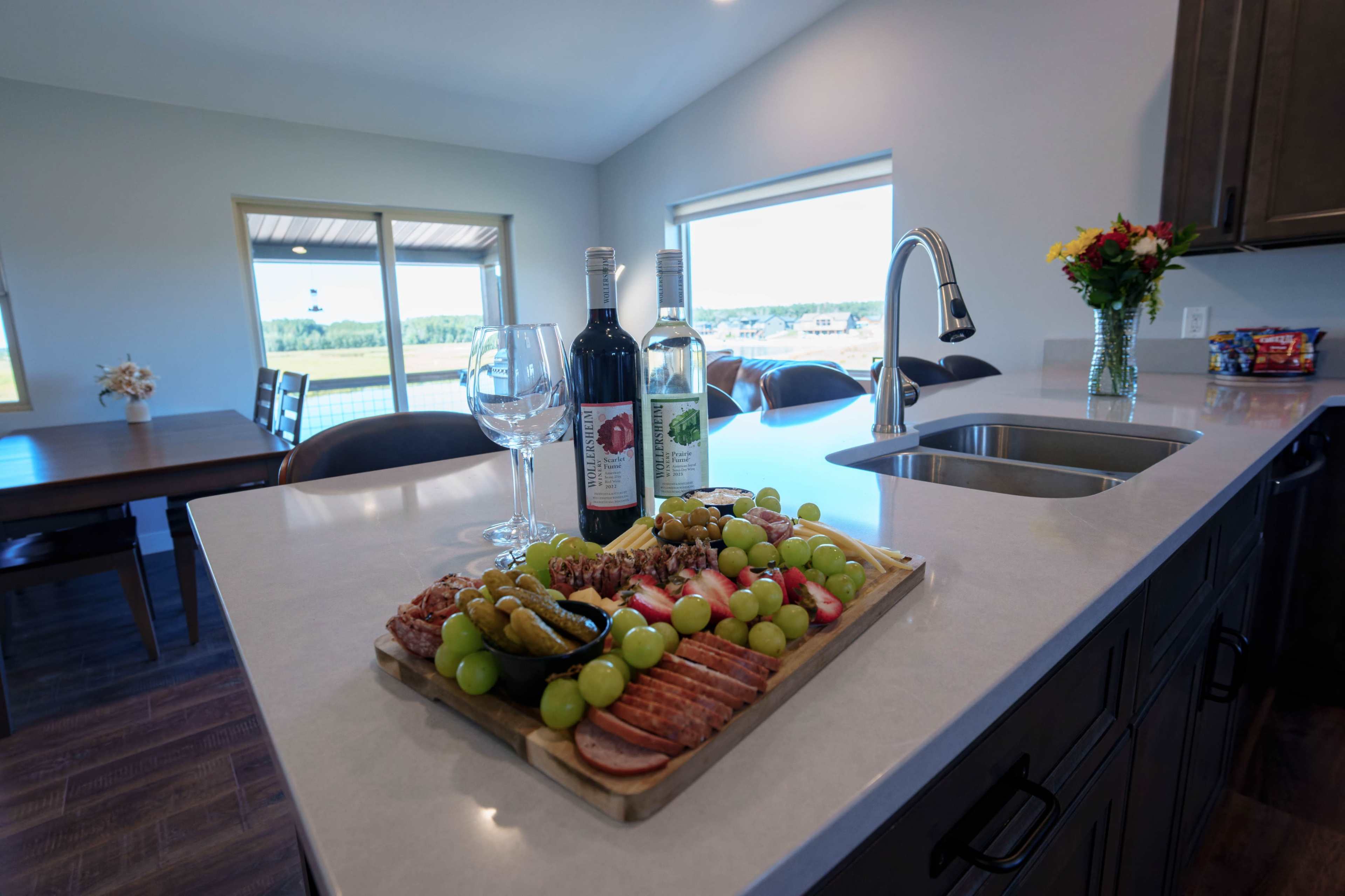 A wooden charcuterie board featuring assorted meats, cheeses, and grapes is displayed on a kitchen counter, alongside bottles of wine and empty wine glasses, with a view of the outdoors visible through large windows.