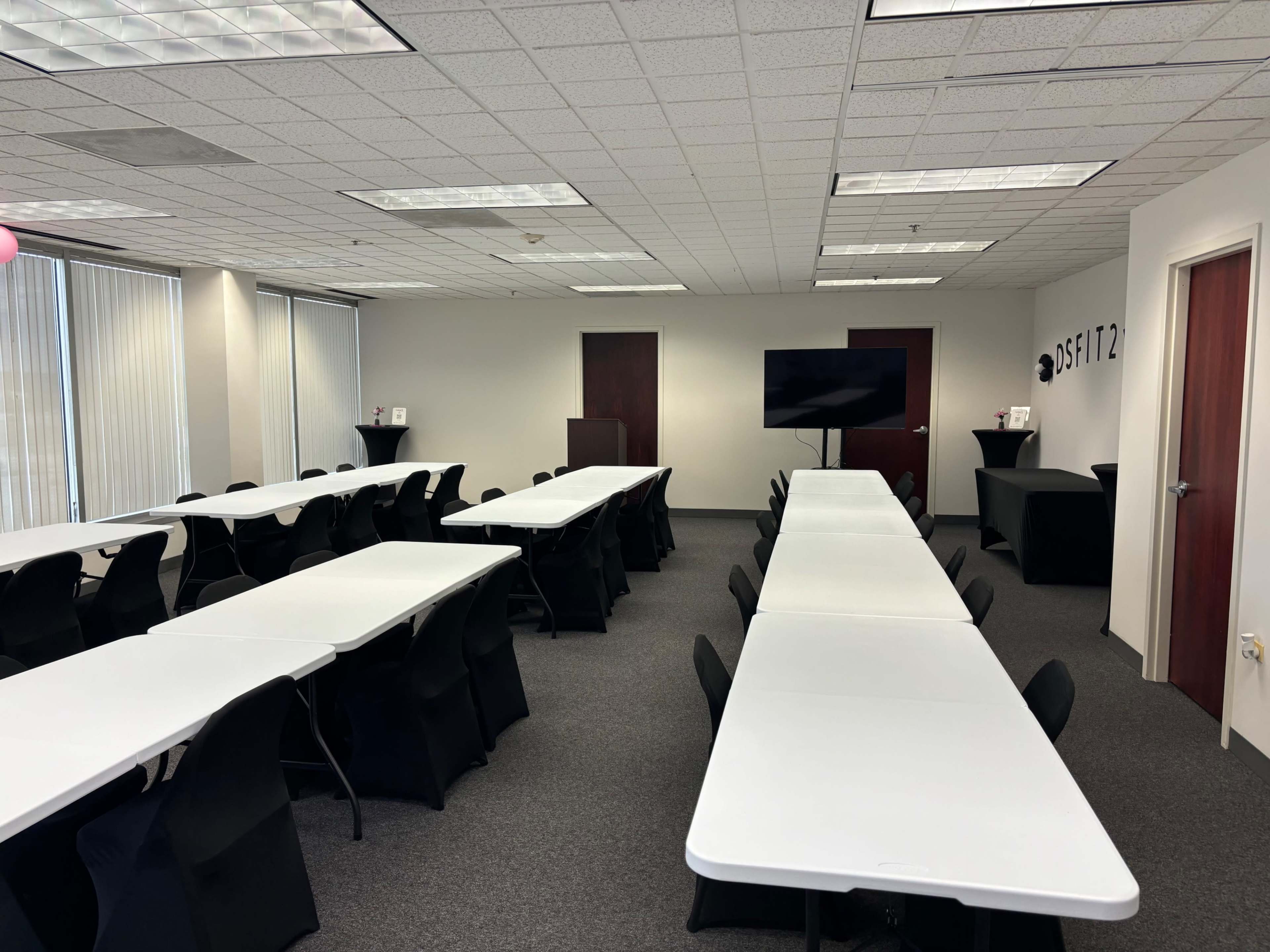 A conference room with several long, white tables arranged in a U-shape and a large screen mounted on the wall at one end.
