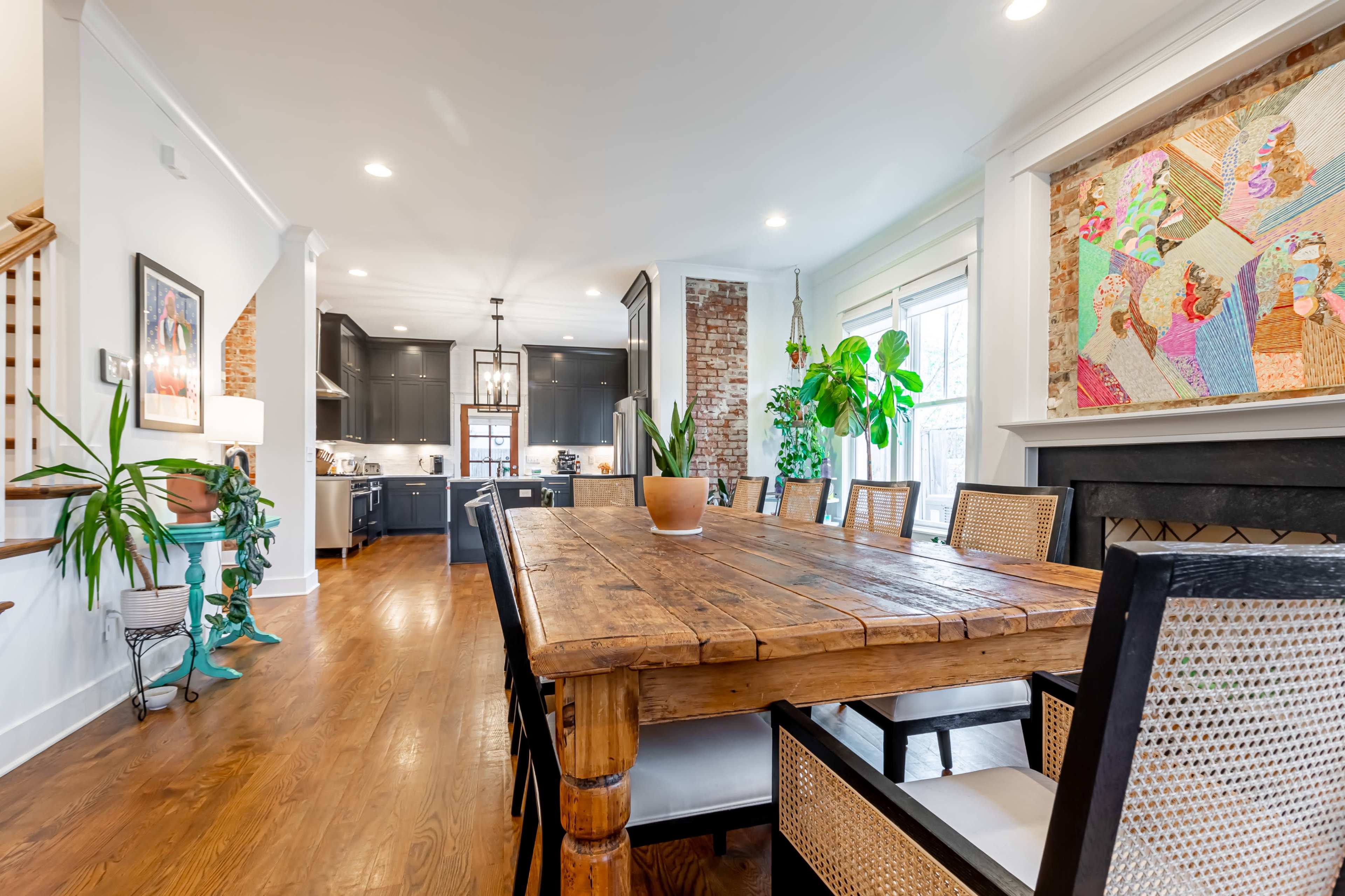 A large wooden dining table is set in a bright room with a kitchen visible in the background and colorful artwork hanging on the wall.