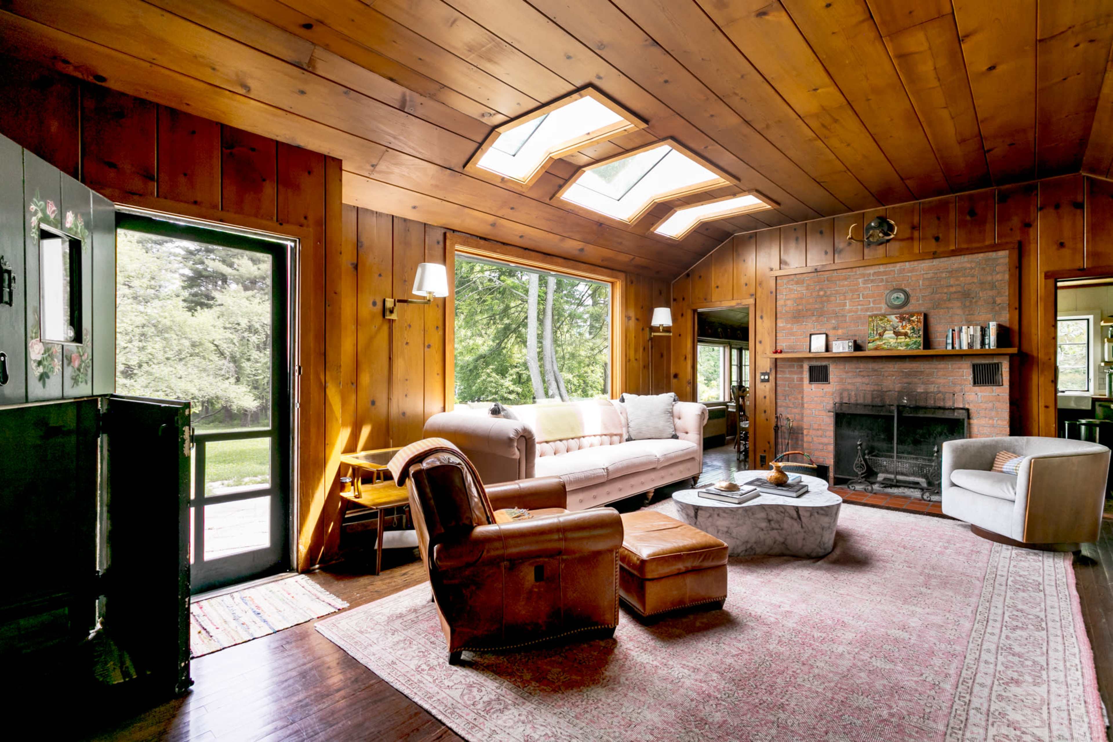 The image features a wooden-paneled living room with a sofa, armchairs, a rug, and a fireplace, illuminated by natural light from skylights and large windows.
