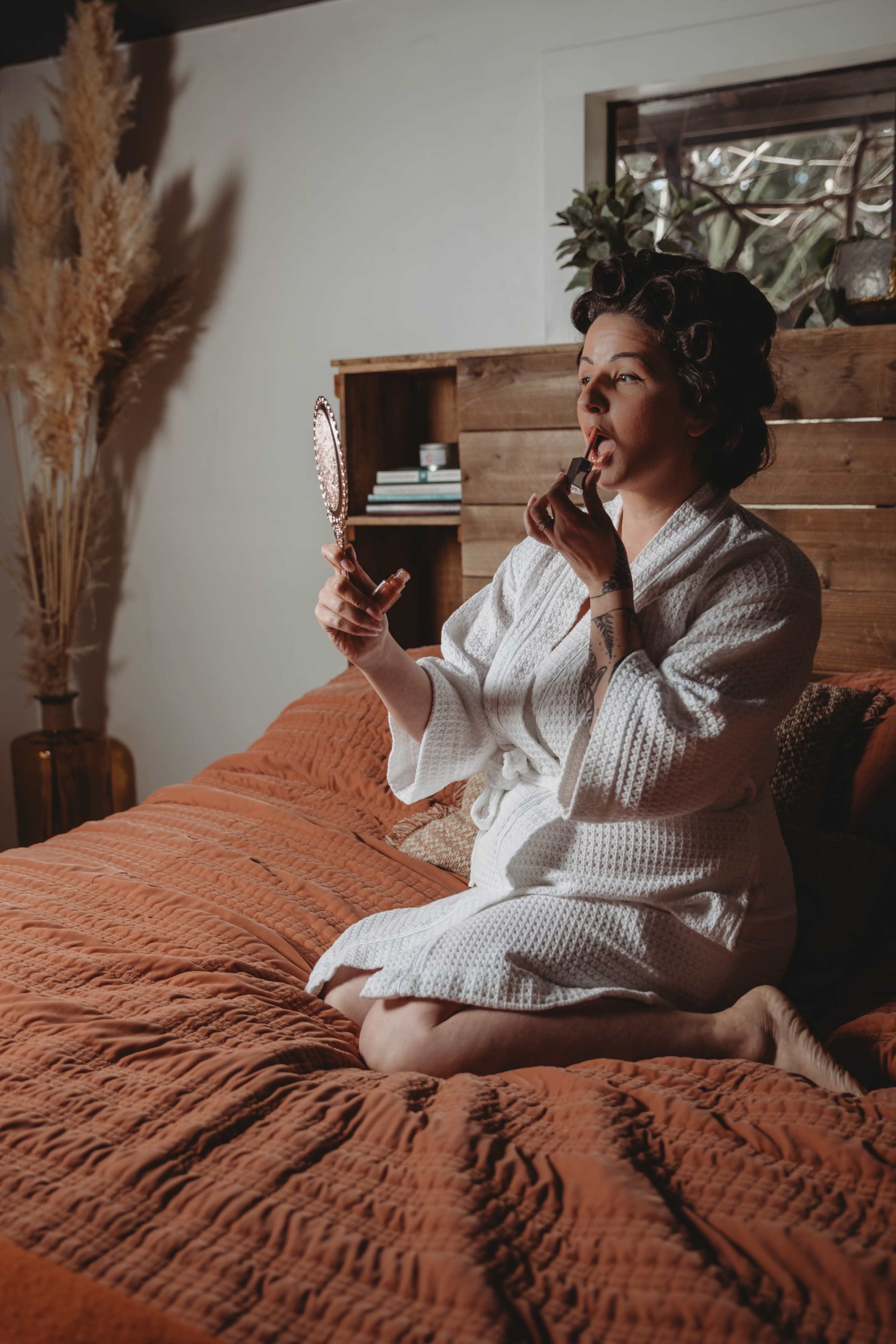 A woman in a white robe sits on a bed with an orange blanket, applying lipstick while looking in a hand mirror.