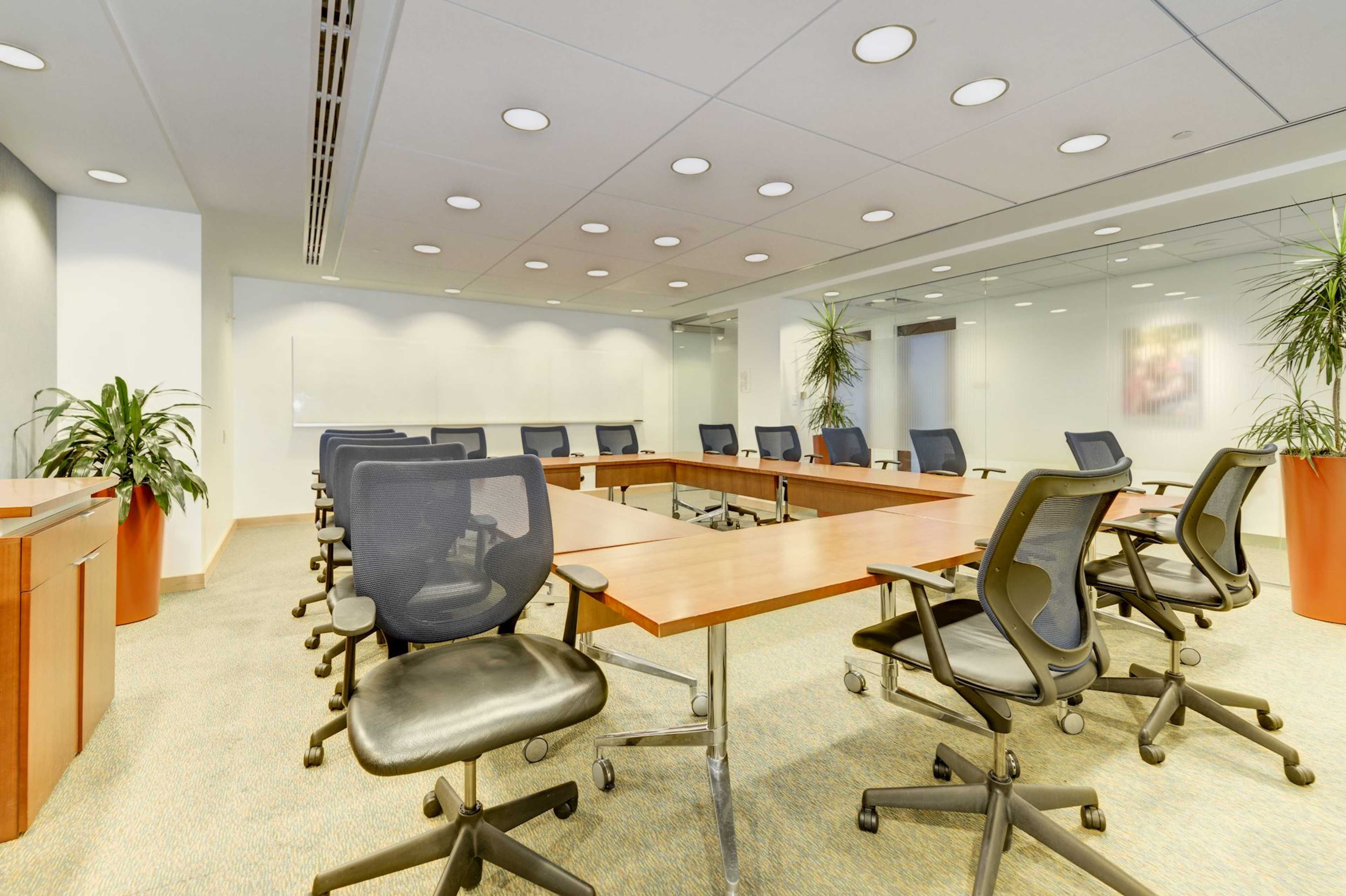 A conference room features a large rectangular table surrounded by rolling office chairs, with plants in decorative pots placed at the corners.