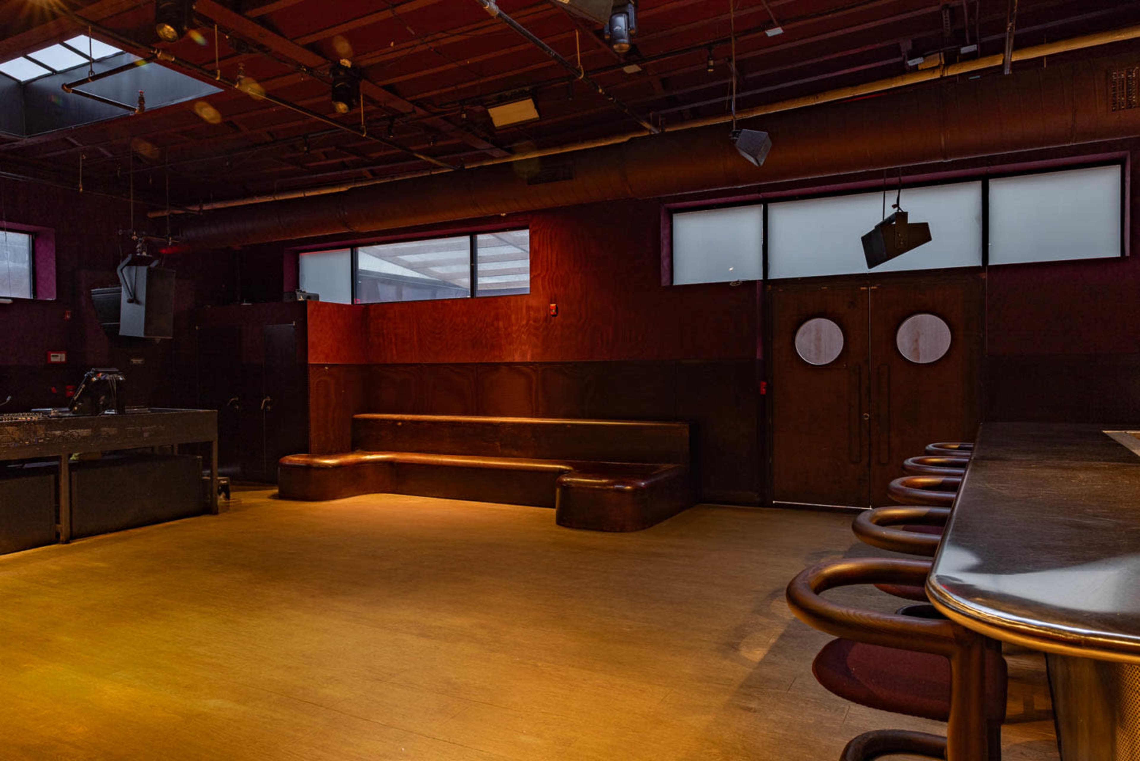 The image shows the interior of a bar with a wooden counter, several round stools, and a small stage area, all set against a backdrop of dark walls and large windows.
