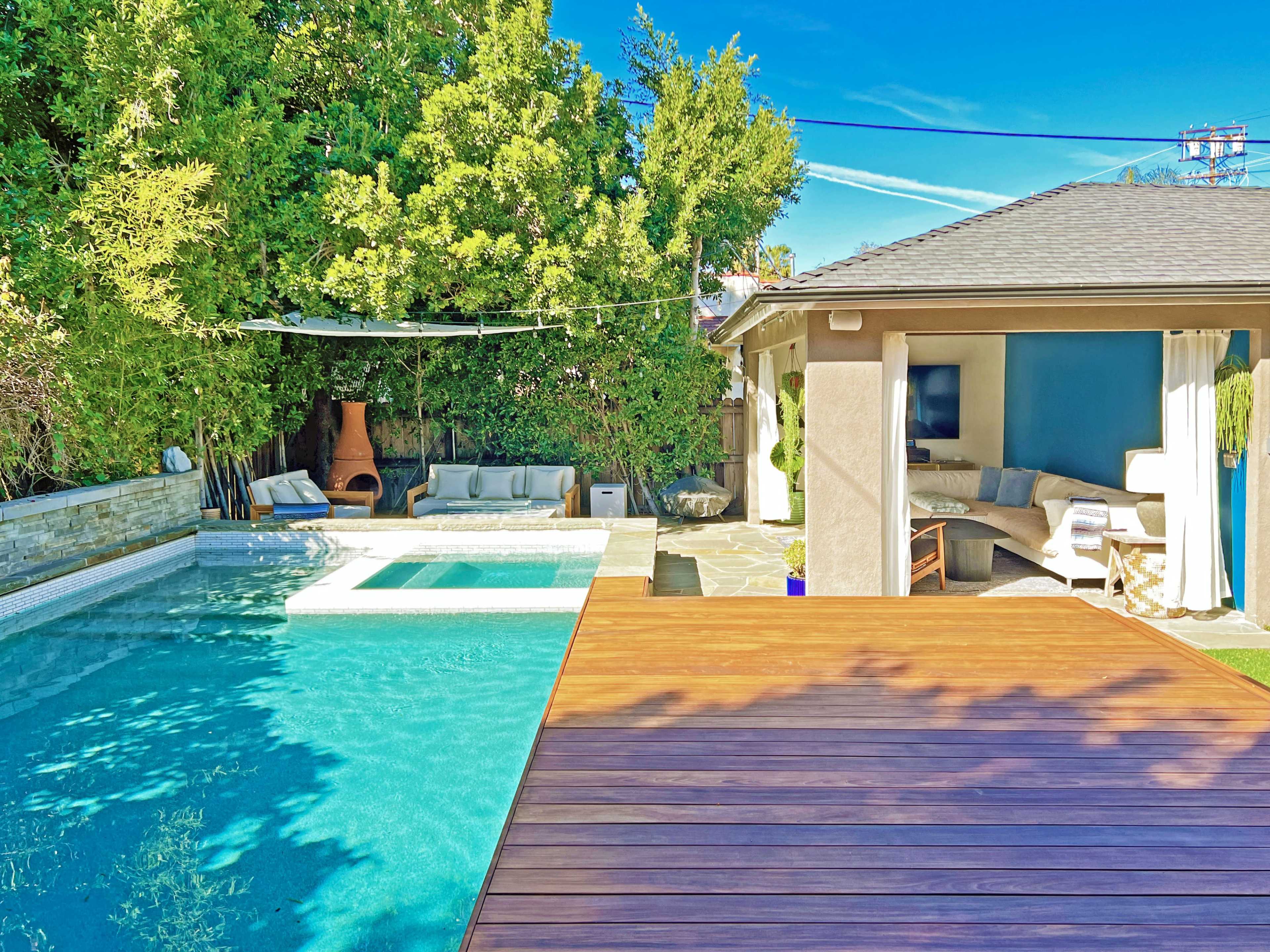 A modern outdoor space featuring a swimming pool, a wooden deck, and a shaded seating area beside lush greenery.