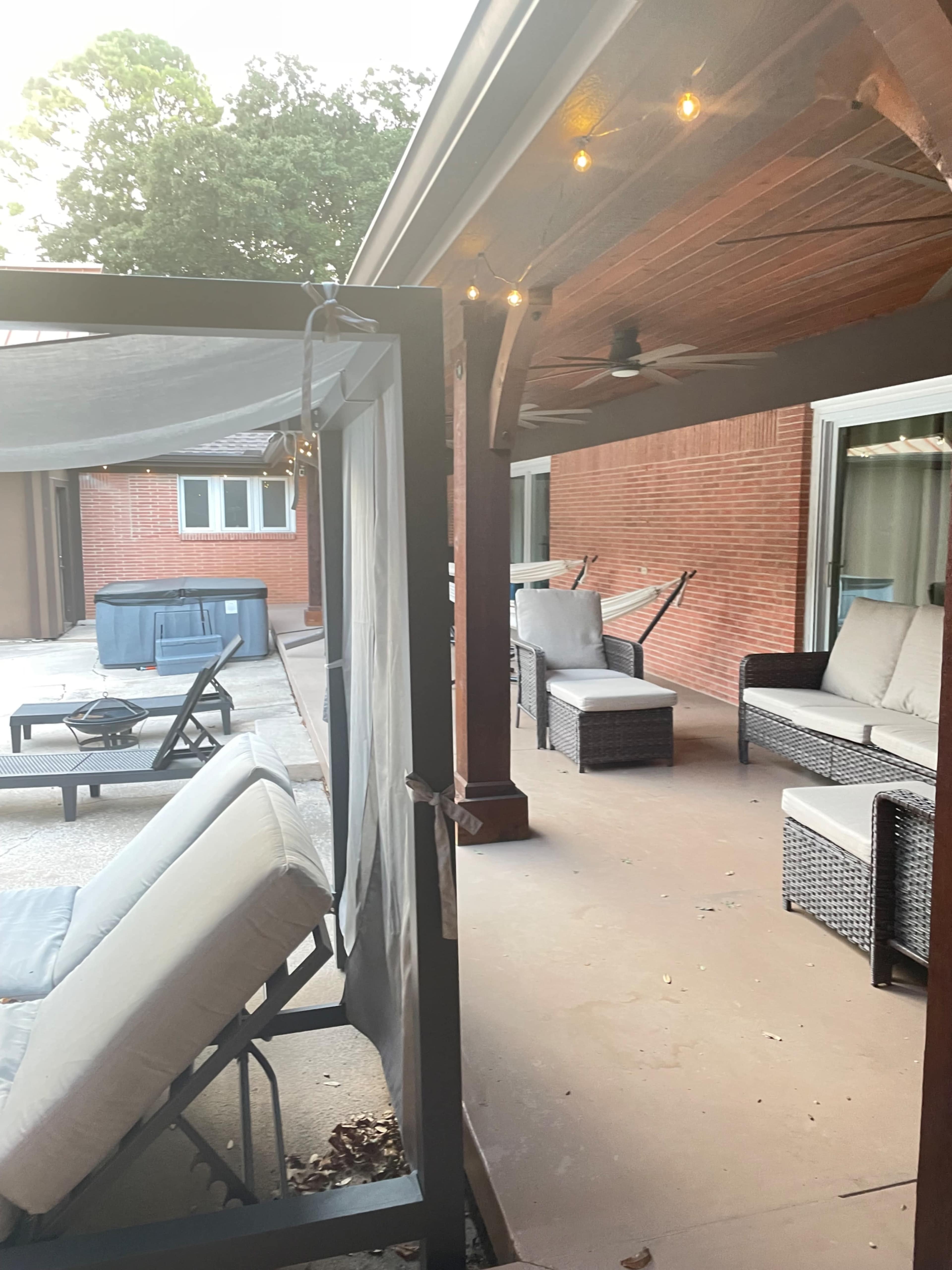The image shows a patio area with outdoor furniture, including a swing and seating arrangements, alongside a hot tub and string lights overhead.