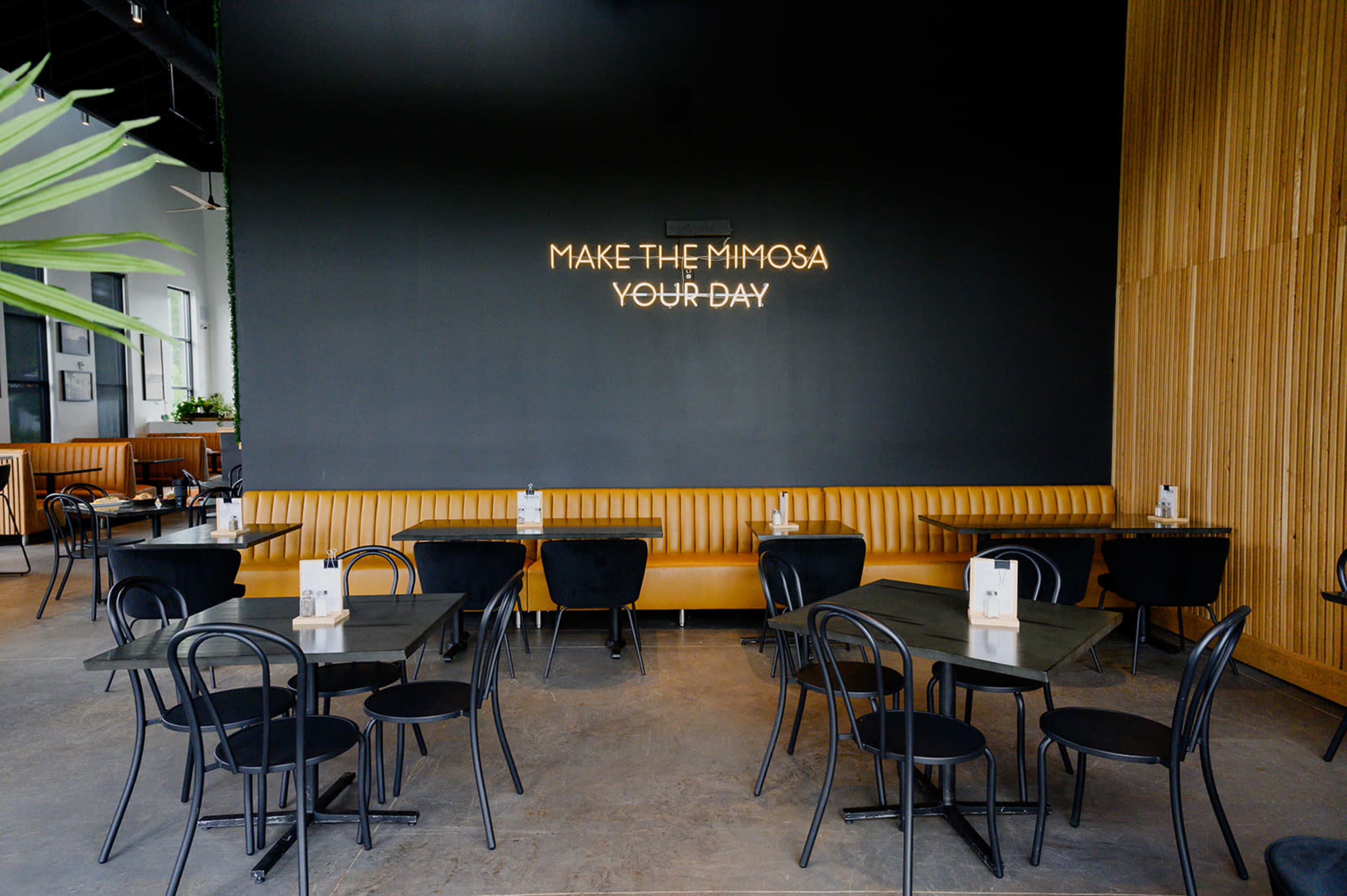 A modern restaurant interior features black chairs and tables arranged around yellow banquette seating, with a neon sign on the wall that reads "MAKE THE MIMOSA YOUR DAY."