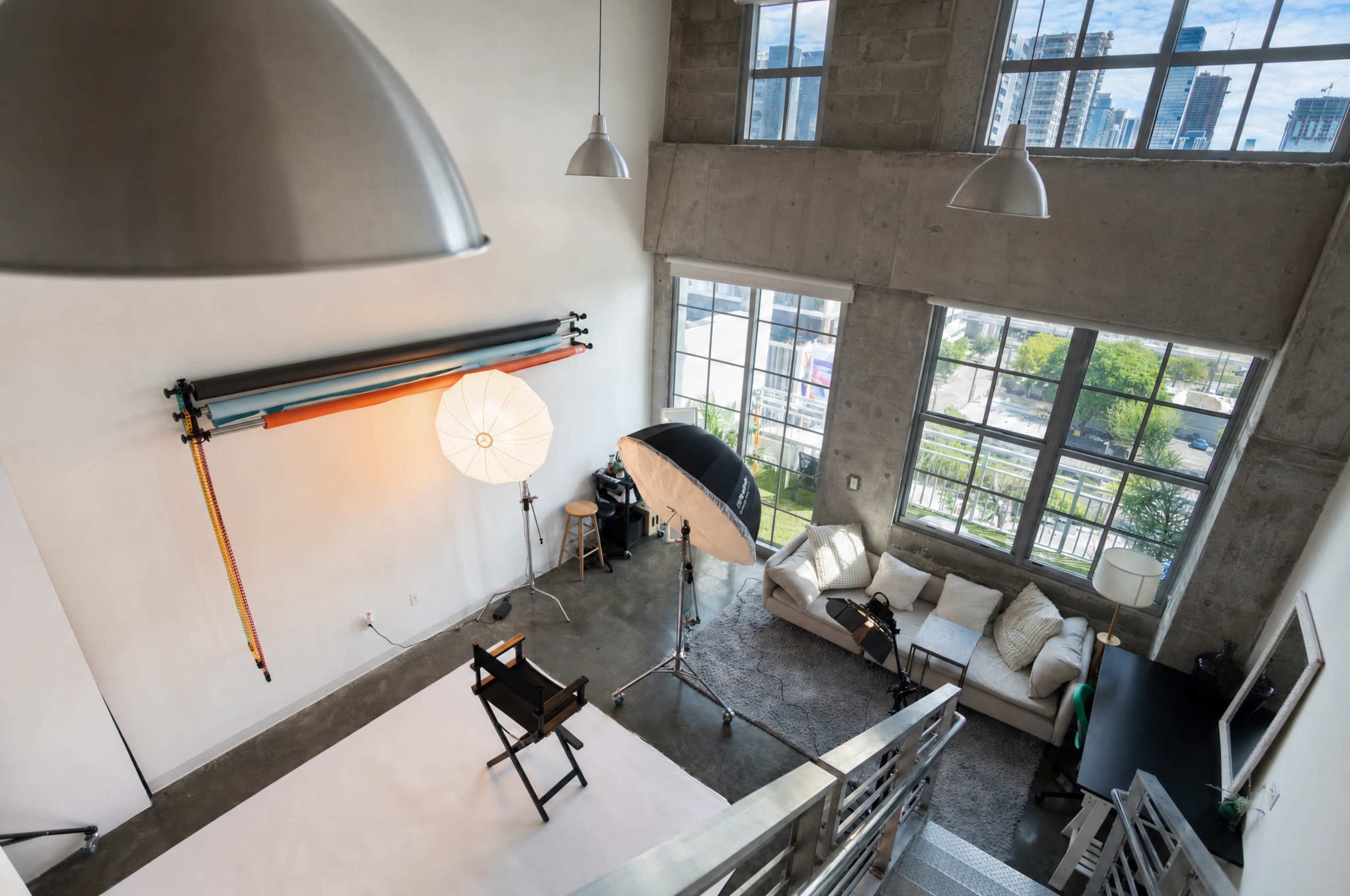 The image shows a spacious photography studio with large windows, a white backdrop, lighting equipment, and a seating area with a couch and a television.