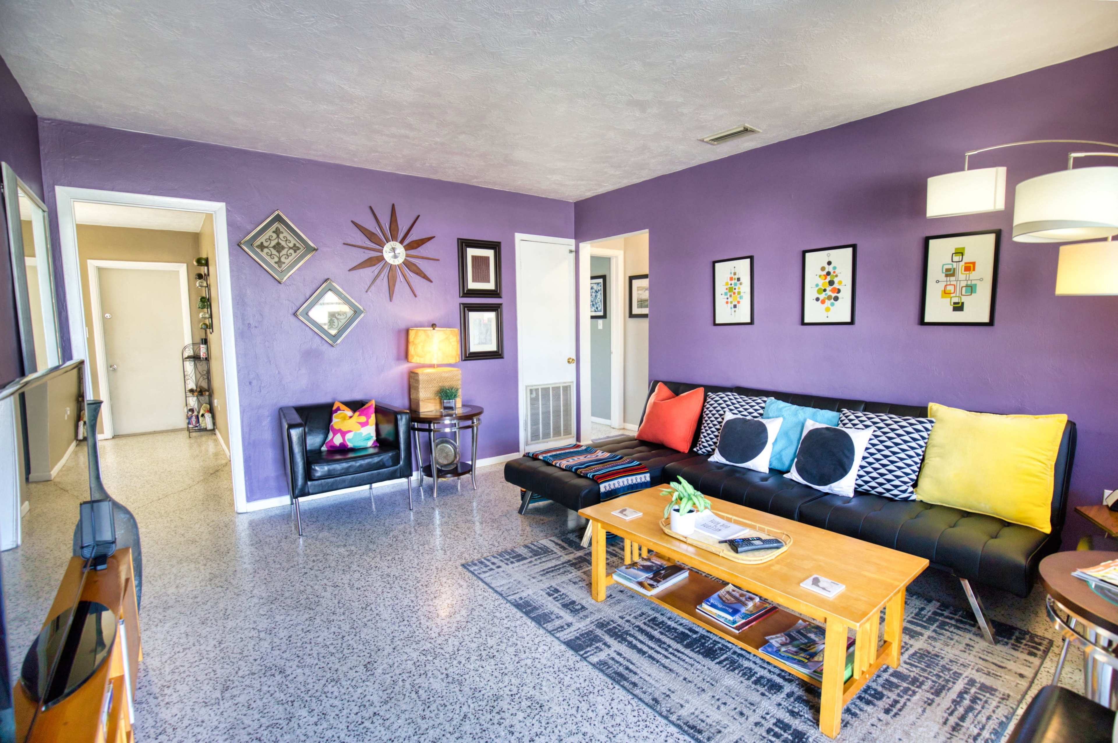 The image shows a modern living room with purple walls, a black sofa, a wooden coffee table, and framed artwork on the walls.