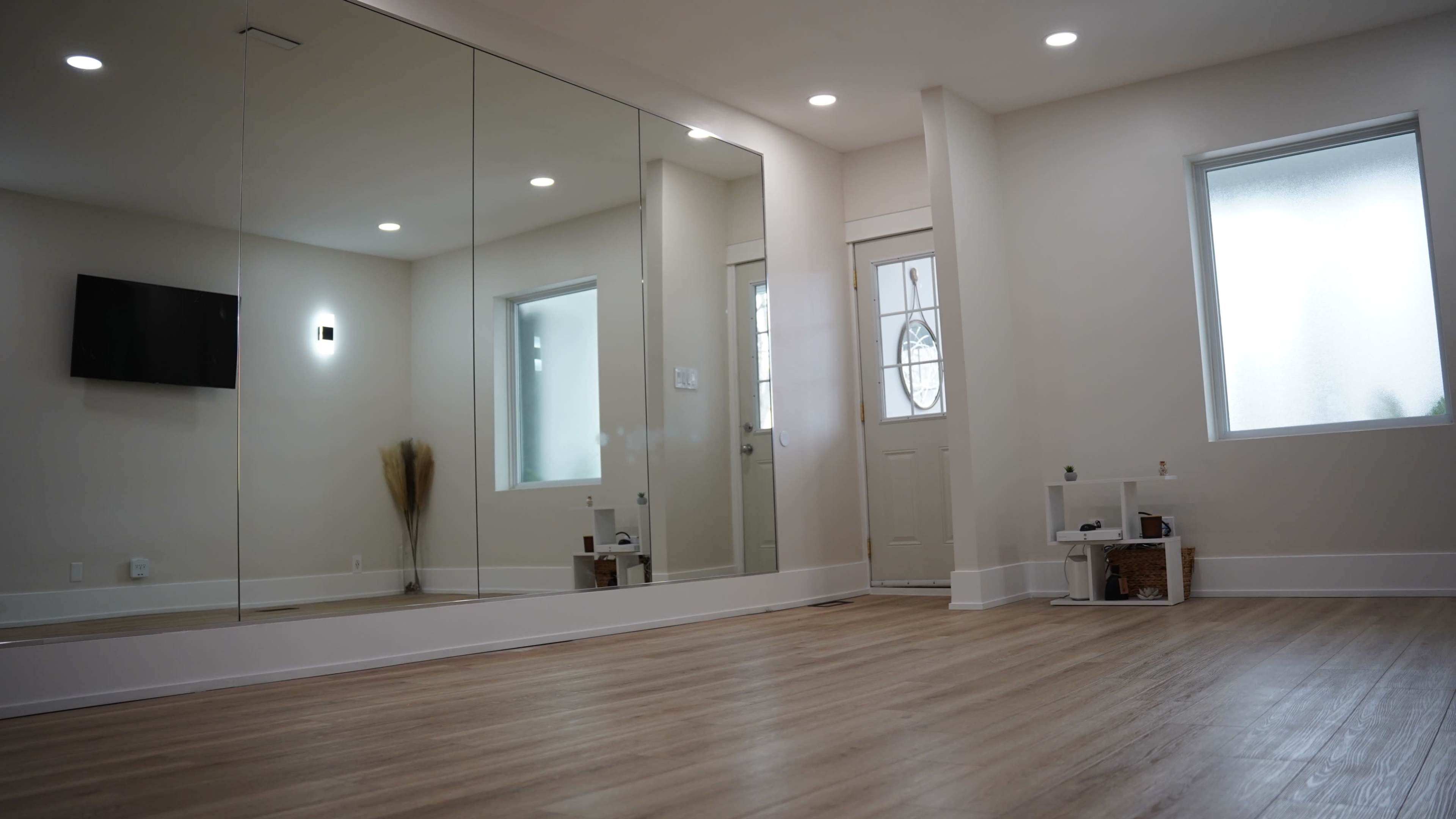 A spacious room with wooden flooring features a large mirror on one wall and a door leading outside.