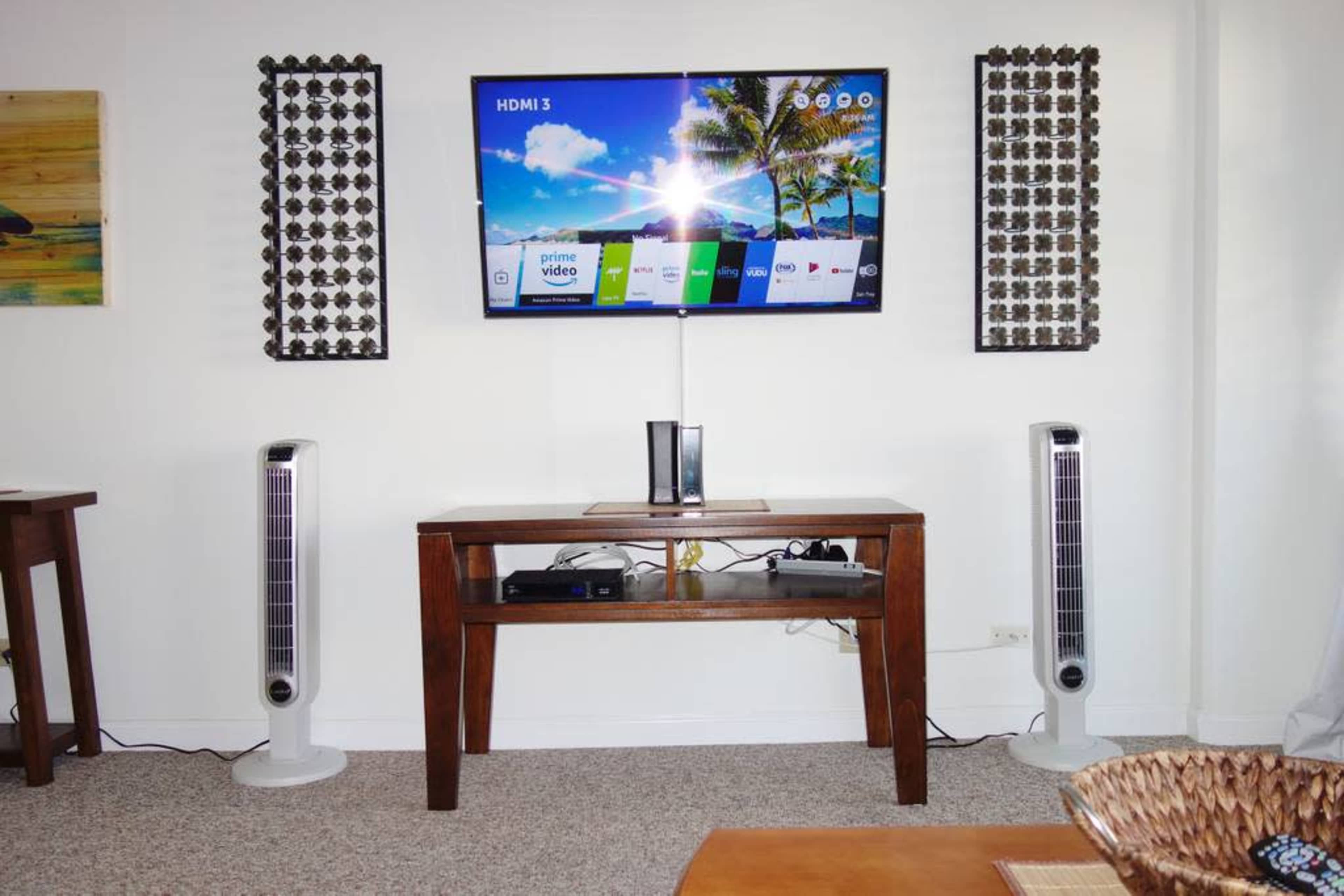 A television is mounted on the wall above a wooden table, flanked by two tall speakers, with a media player and cables visible underneath the table.