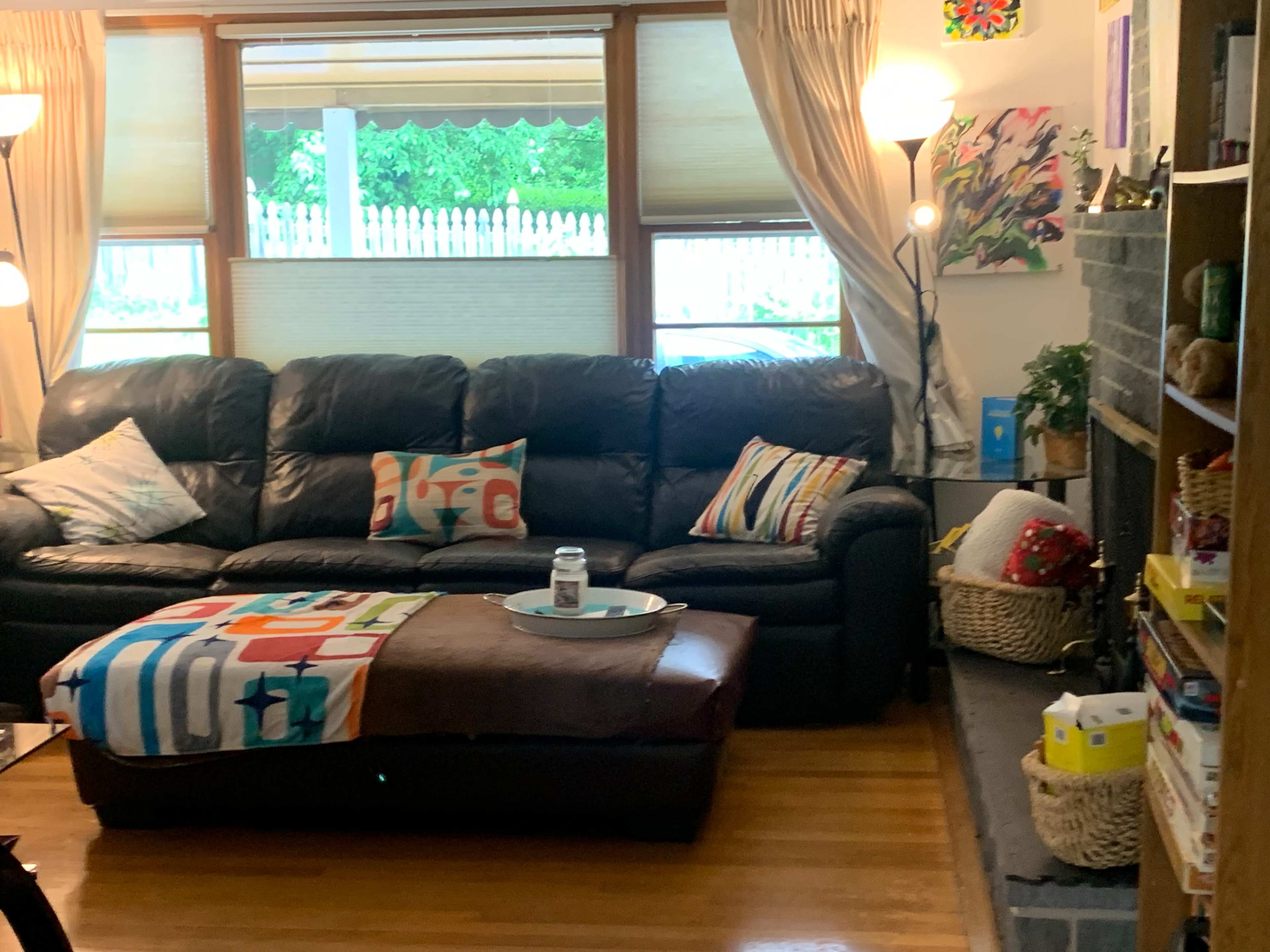 The image shows a living room featuring a black leather sofa with colorful throw pillows, a coffee table in front, and a bookshelf filled with various items.