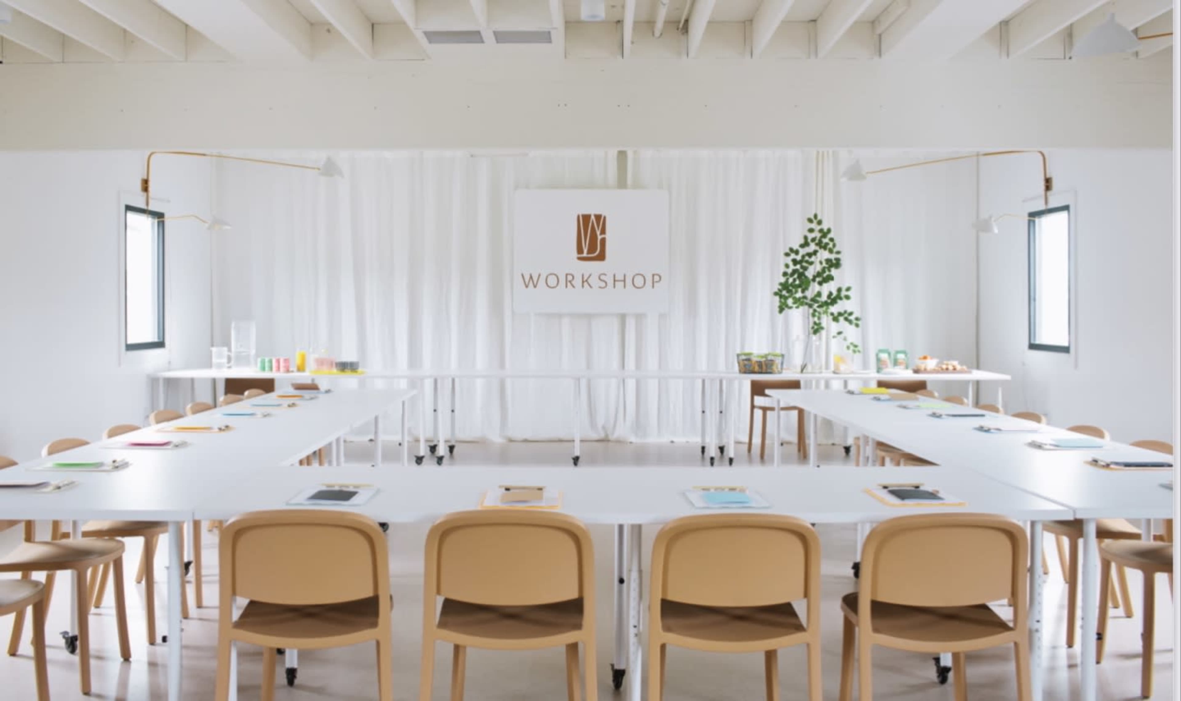 A bright, modern workshop space features a long table set with materials, surrounded by light-colored chairs and green plants.