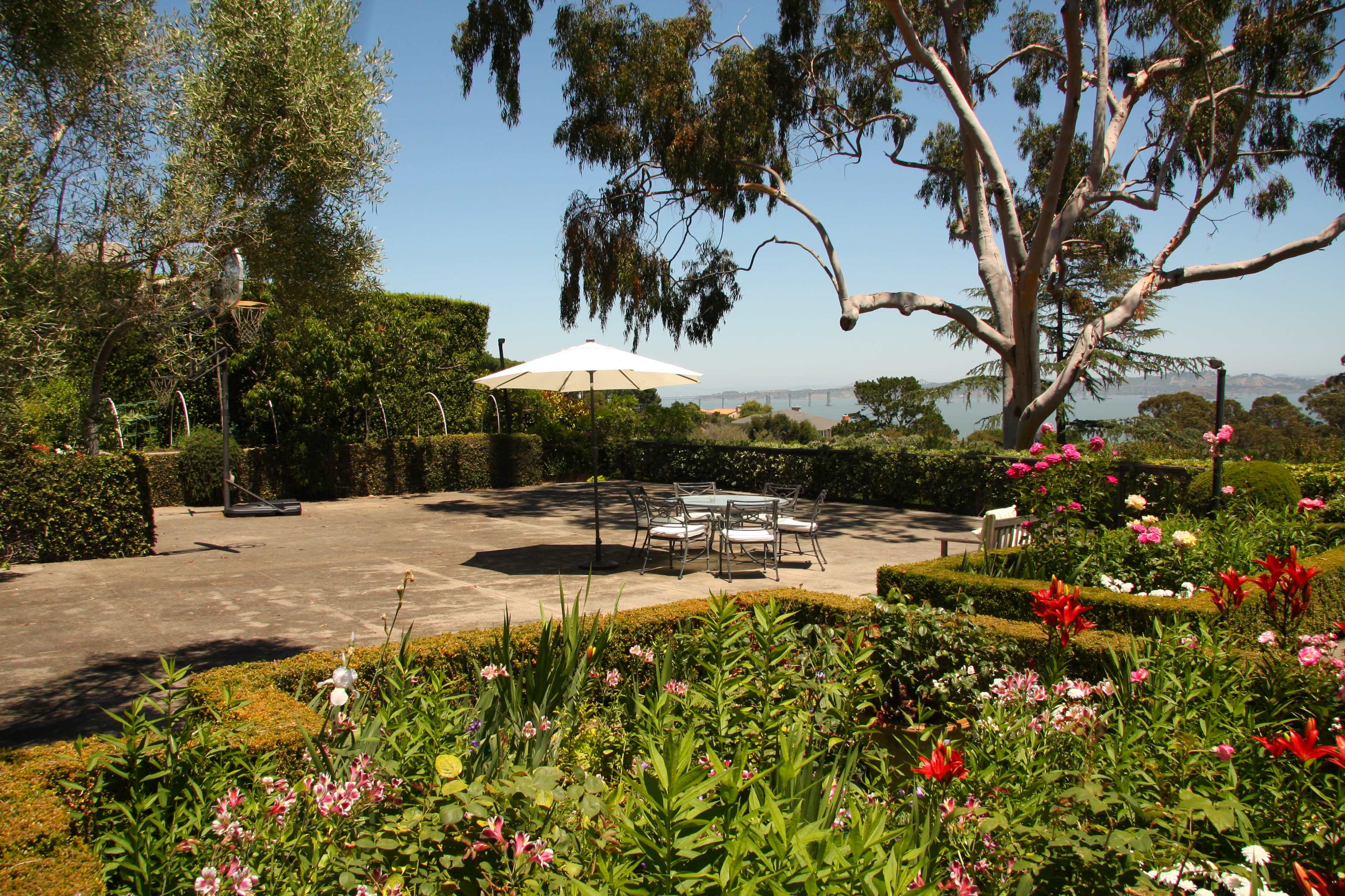 The image shows a landscaped garden with a stone patio, outdoor seating, and vibrant flowers, overlooking a distant water view.