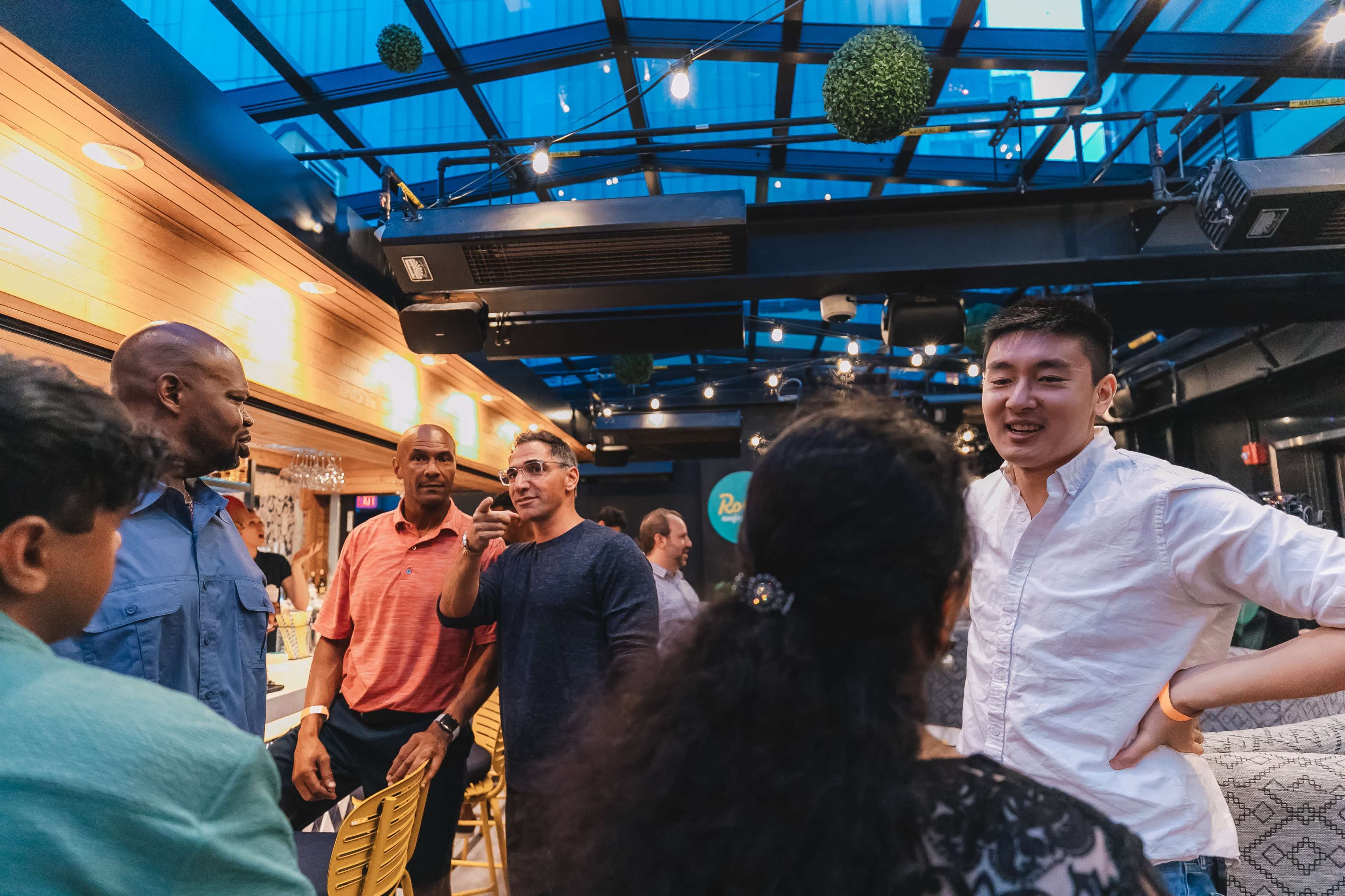 A group of people are engaged in conversation inside a well-lit venue with a glass ceiling and hanging greenery.