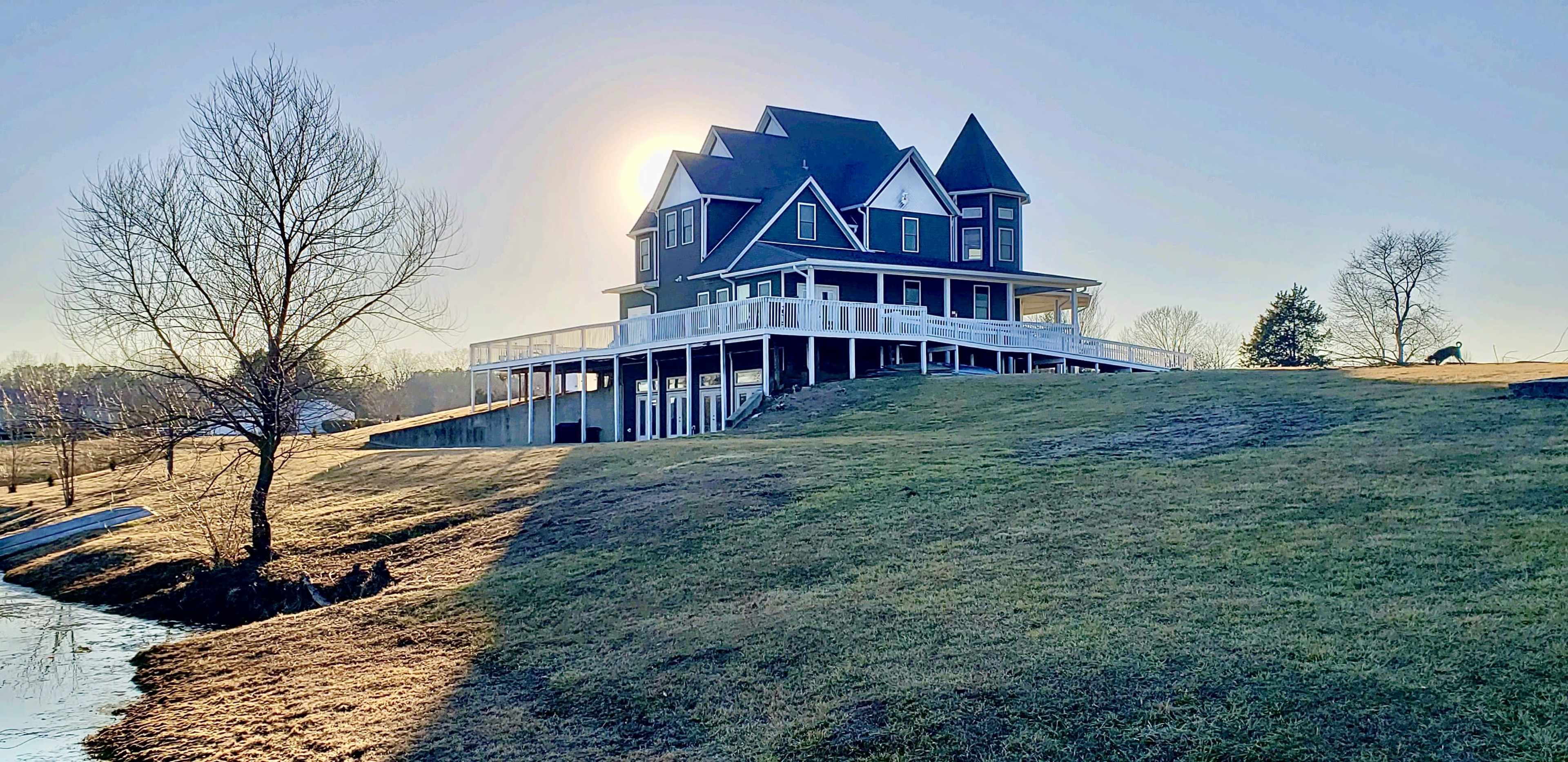 A large house with a wraparound porch sits on a grassy hill, overlooking a body of water under a clear sky.