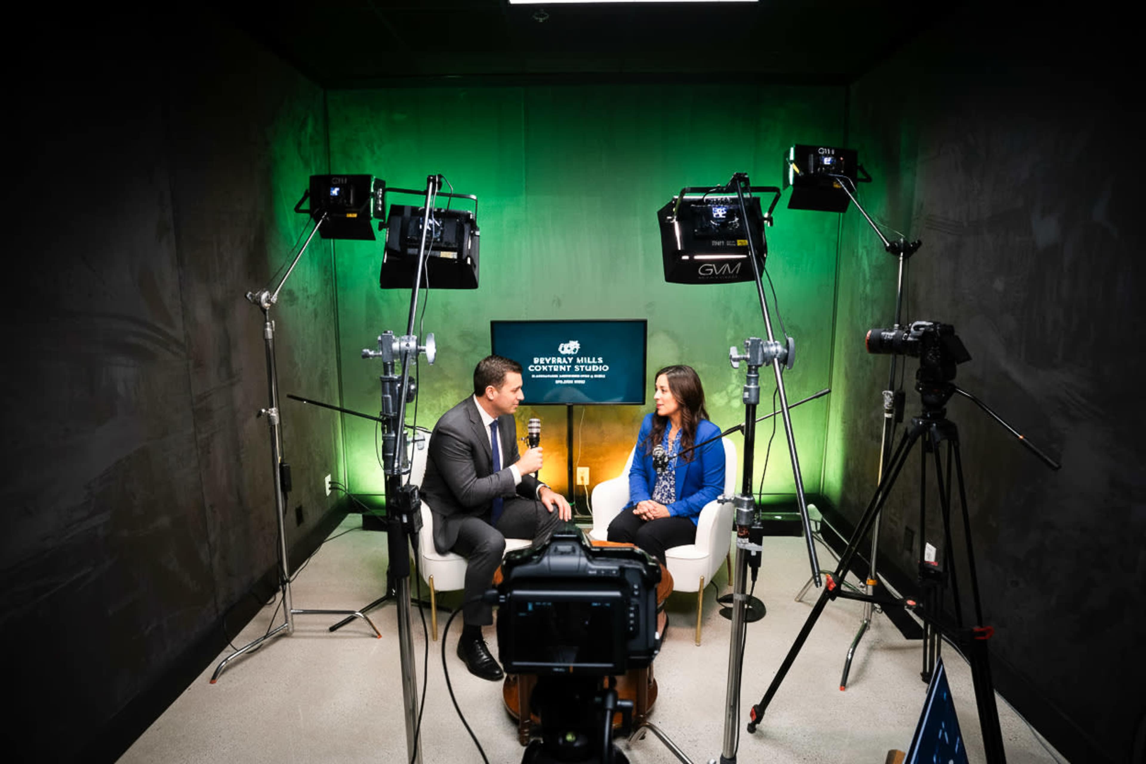 A man and a woman are seated in a studio setting, with multiple cameras and lights positioned around them, as they engage in conversation.
